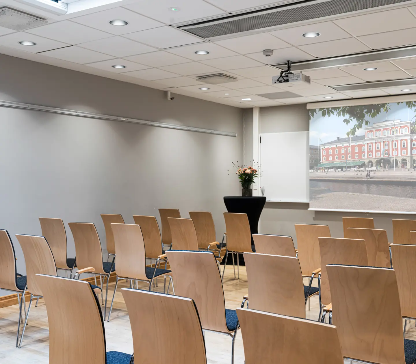 Modern conference room setup with rows of wooden chairs facing a projector screen displaying an exterior view of a building. Ideal for business meetings or presentations.