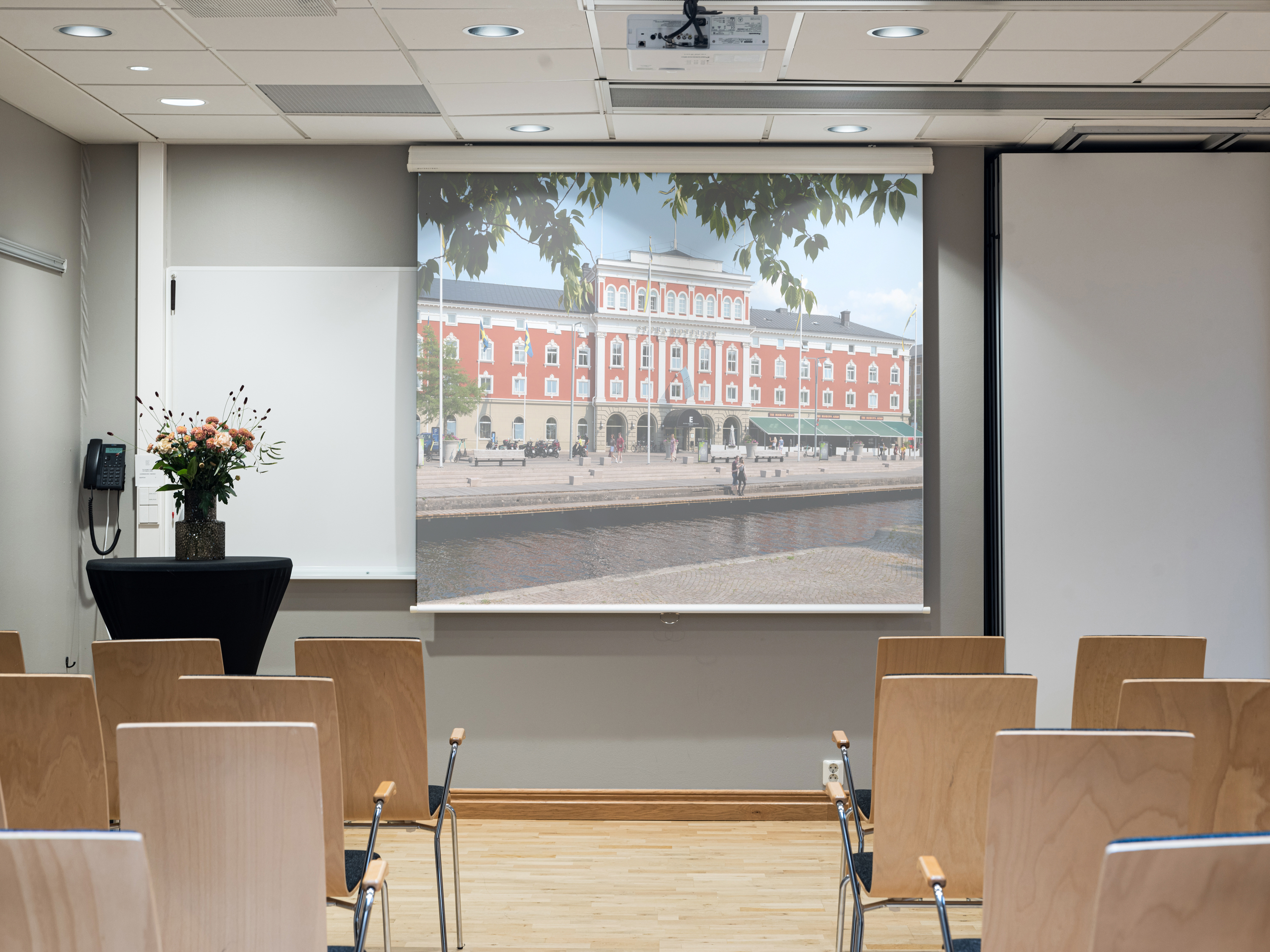 A conference room with wooden chairs facing a screen displaying an image of a red brick building by a canal. A vase of flowers is on a small table to the side, and there is a whiteboard and a telephone on the wall.