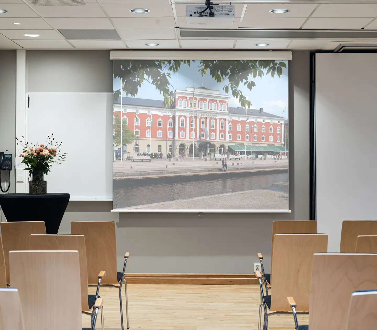 A conference room with wooden chairs facing a screen displaying an image of a red brick building by a canal. A vase of flowers is on a small table to the side, and there is a whiteboard and a telephone on the wall.