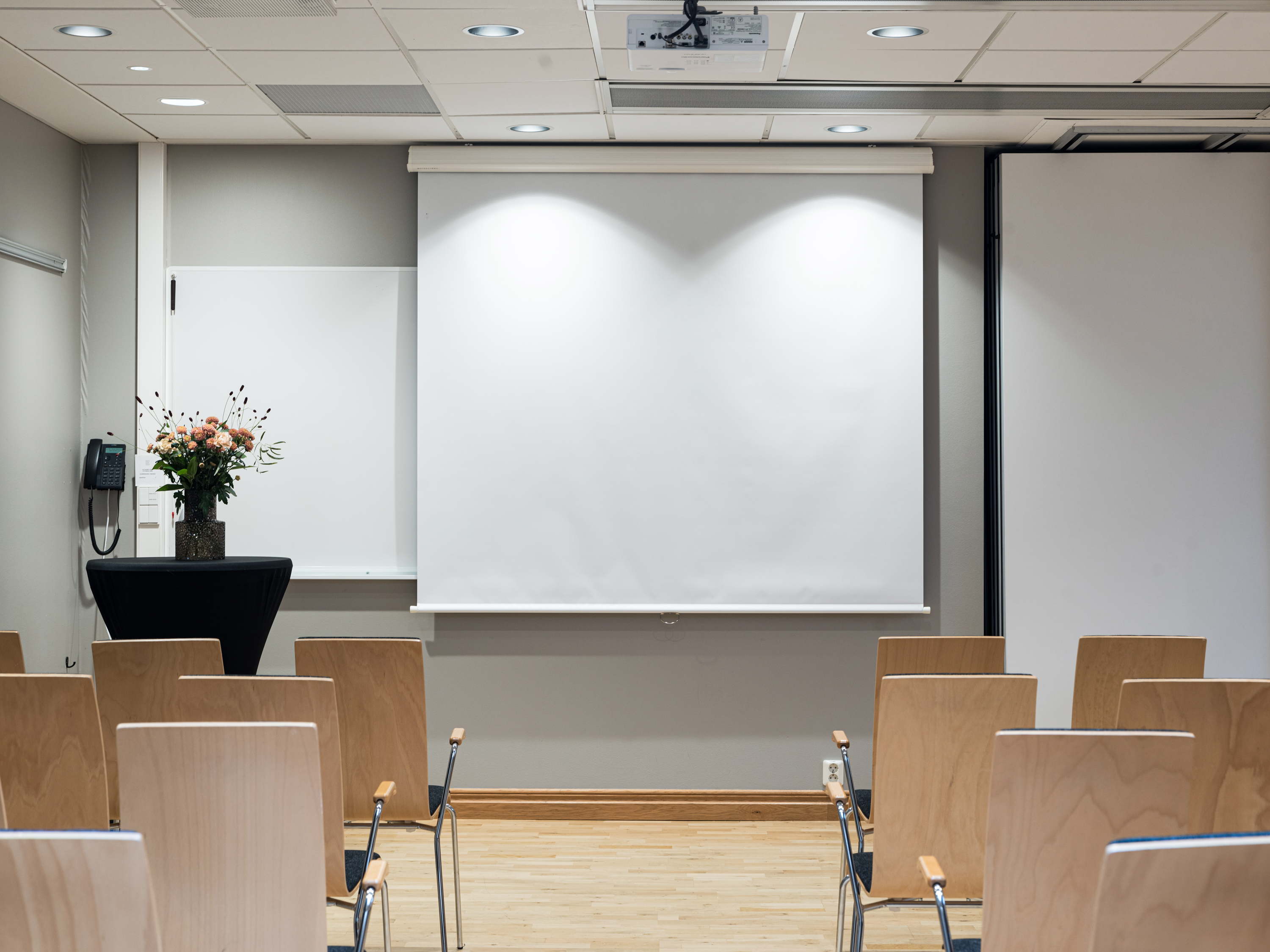 Empty conference room with rows of wooden chairs facing a white projection screen and a floral arrangement on a table.