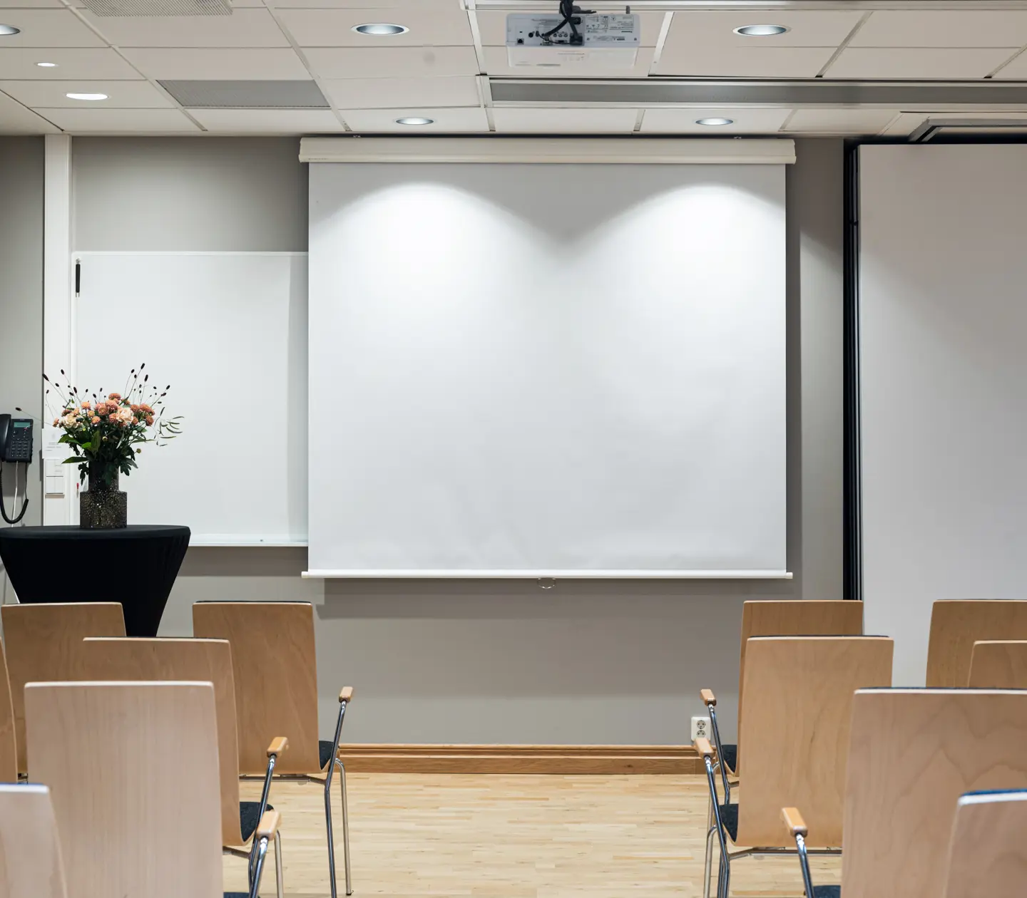 Empty conference room with rows of wooden chairs facing a white projection screen and a floral arrangement on a table.