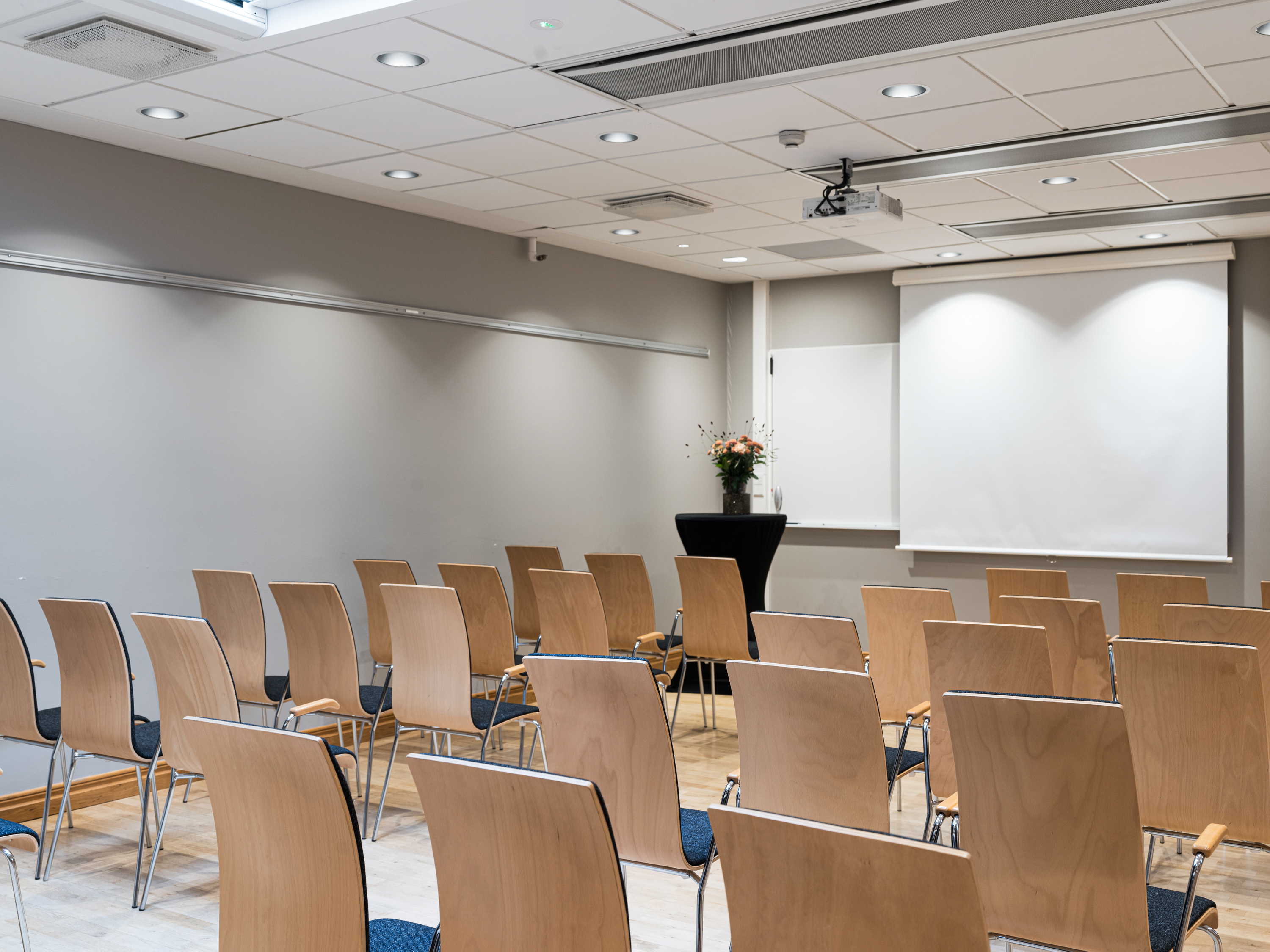 Empty conference room with wooden chairs facing a white projection screen, gray walls, and bright overhead lighting.