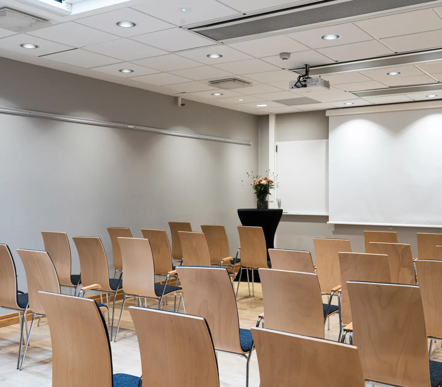 Empty conference room with wooden chairs facing a white projection screen, gray walls, and bright overhead lighting.