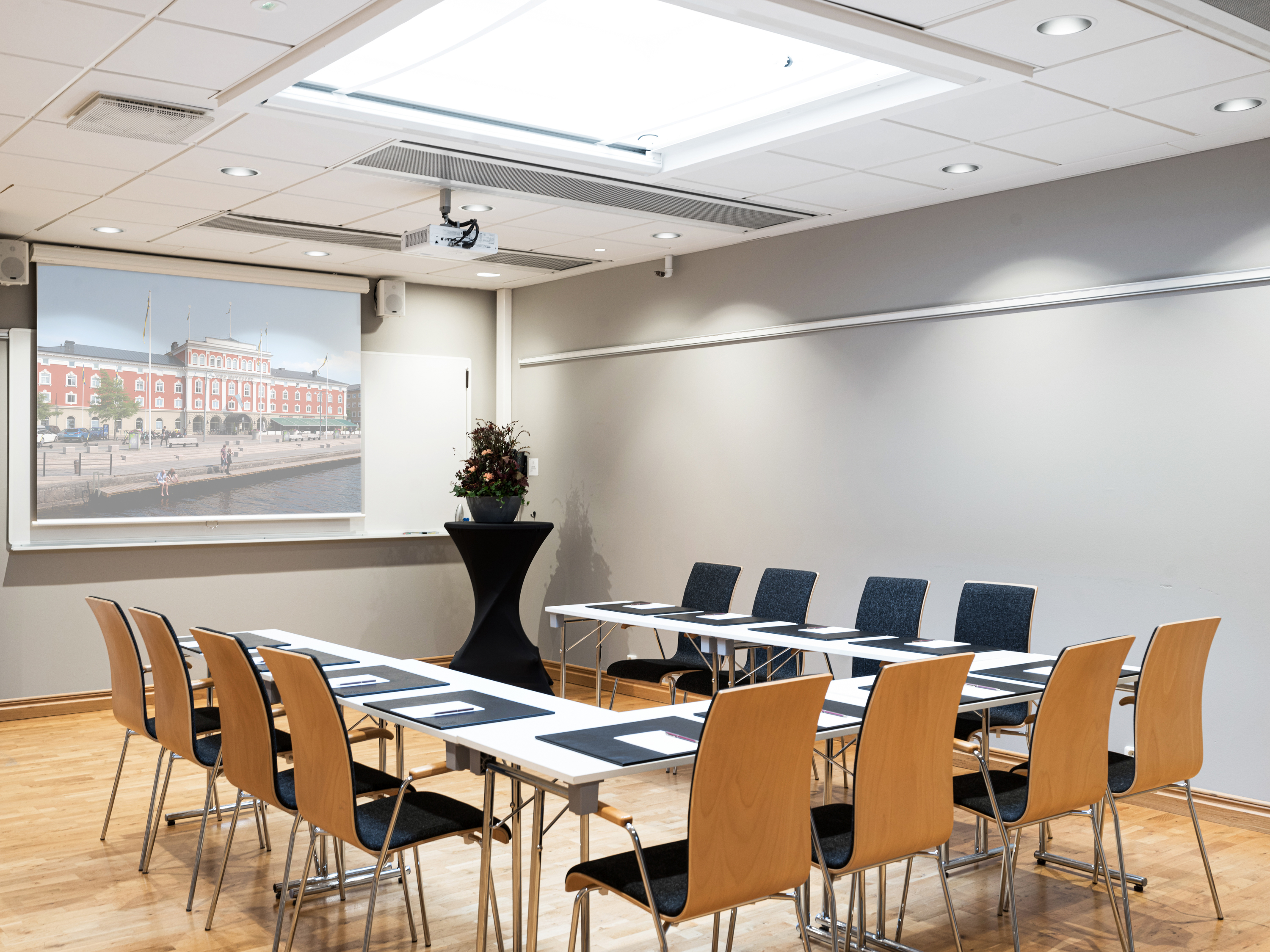 Meeting room with wooden chairs, rectangular tables arranged in a U-shape, a projector screen displaying a cityscape, and a small round table with a flower arrangement.