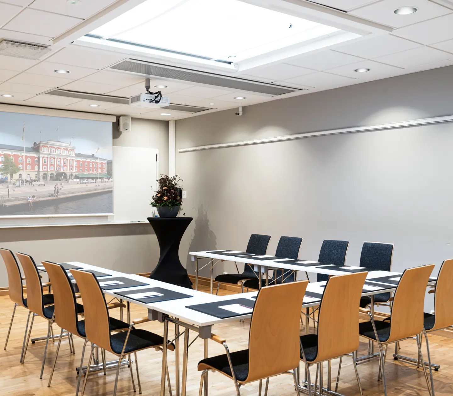 Meeting room with wooden chairs, rectangular tables arranged in a U-shape, a projector screen displaying a cityscape, and a small round table with a flower arrangement.