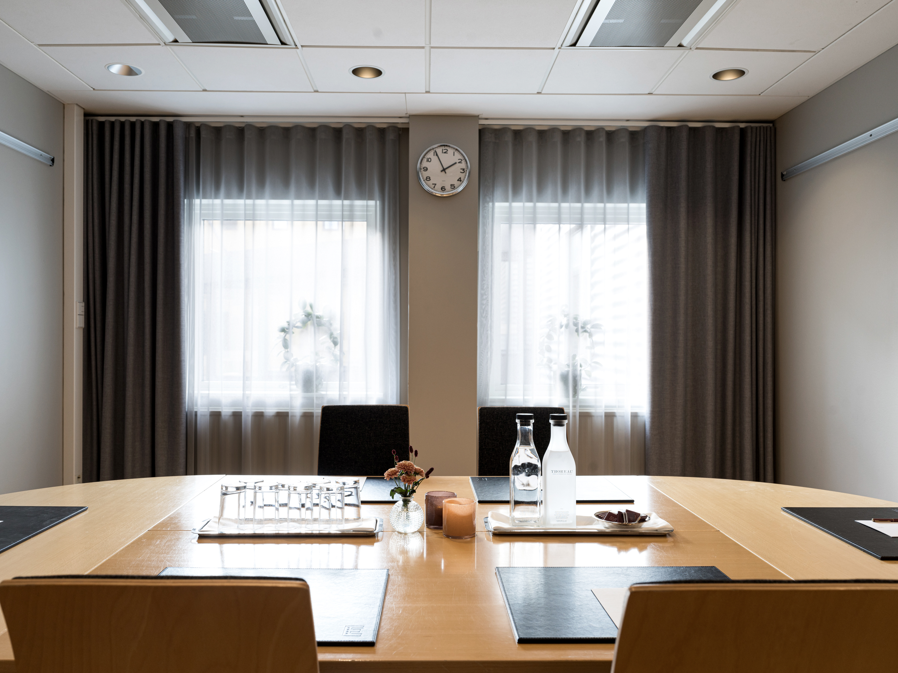Modern conference room with a wooden table set for a meeting, featuring water bottles, a clock on the wall, and natural light coming through curtained windows.