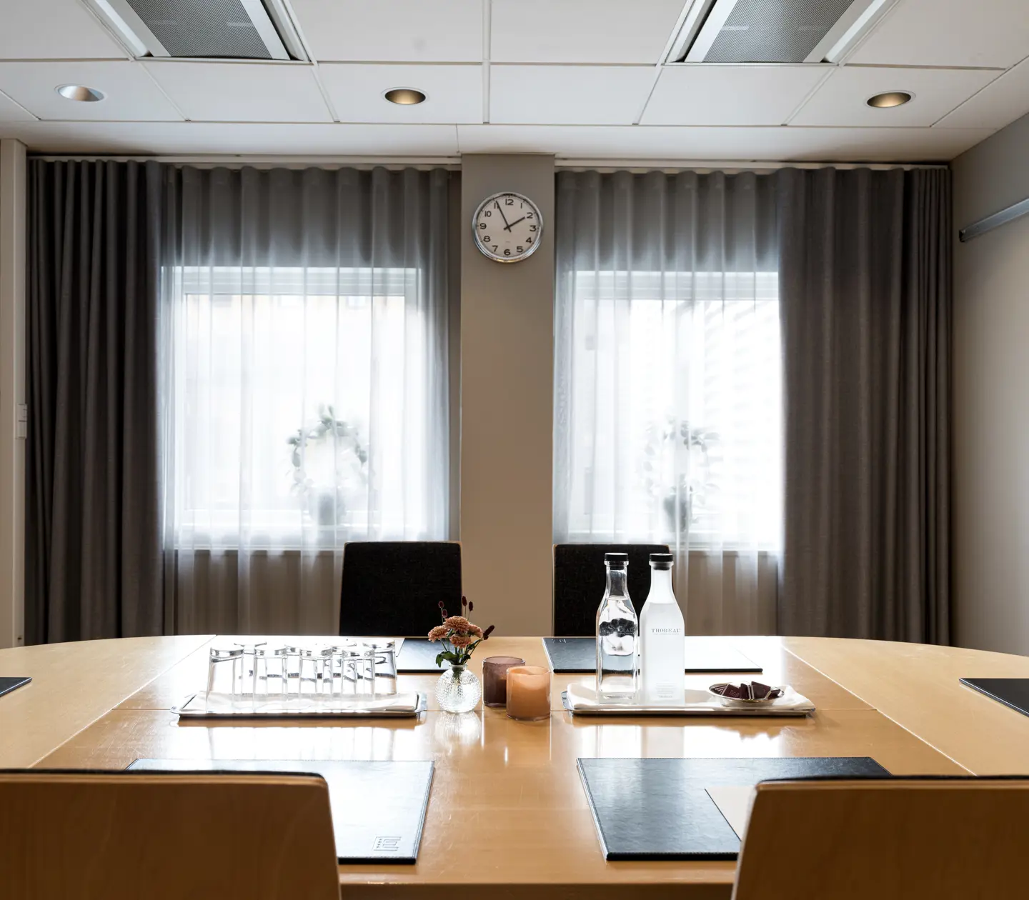 Modern conference room with a wooden table set for a meeting, featuring water bottles, a clock on the wall, and natural light coming through curtained windows.