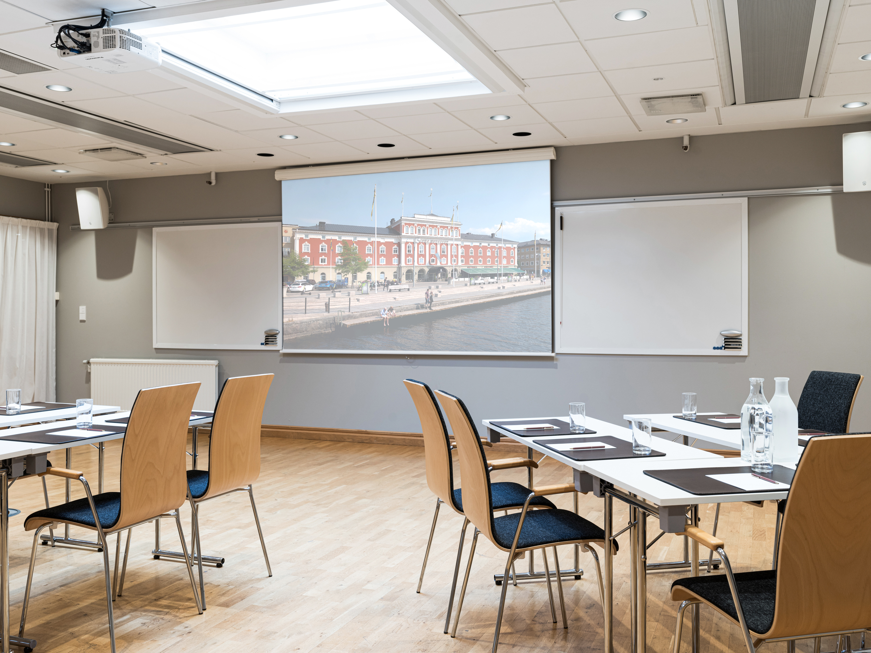 Modern conference room with wooden chairs and white tables, featuring a large screen displaying an exterior building view, wooden flooring, and ample natural light from the ceiling.
