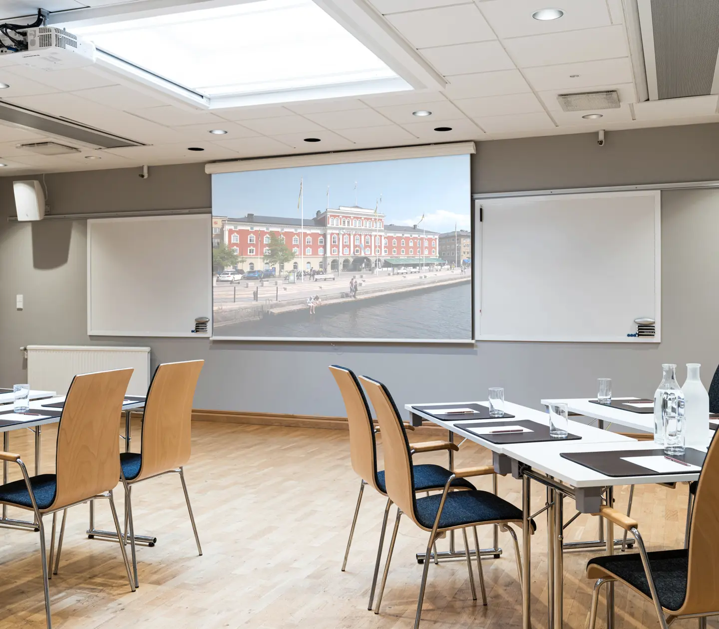 Modern conference room with wooden chairs and white tables, featuring a large screen displaying an exterior building view, wooden flooring, and ample natural light from the ceiling.