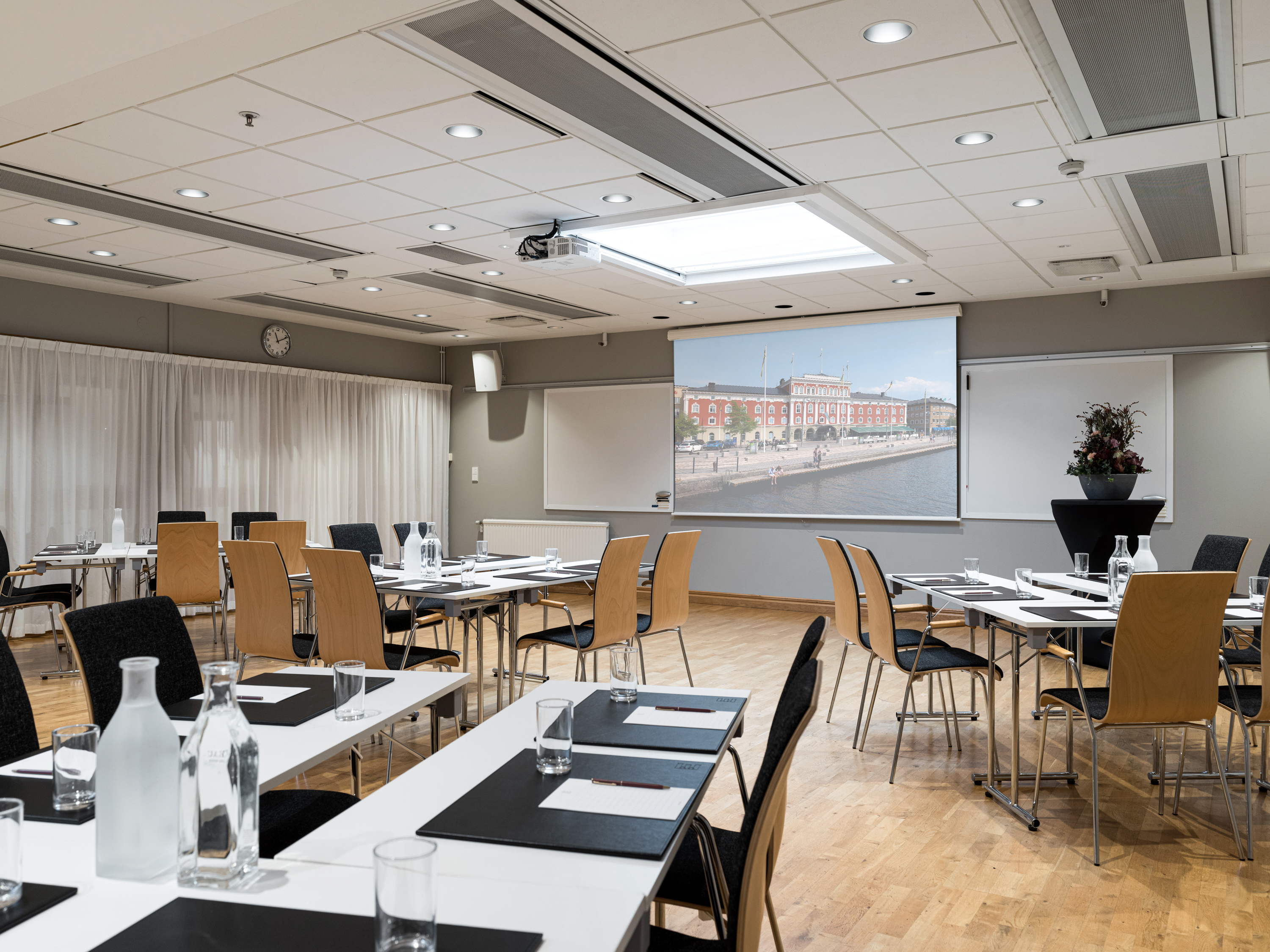 Spacious conference room setup with tables and chairs, featuring a large screen displaying a cityscape. The room has modern decor, a wooden floor, and is well-lit, ideal for meetings or presentations.
