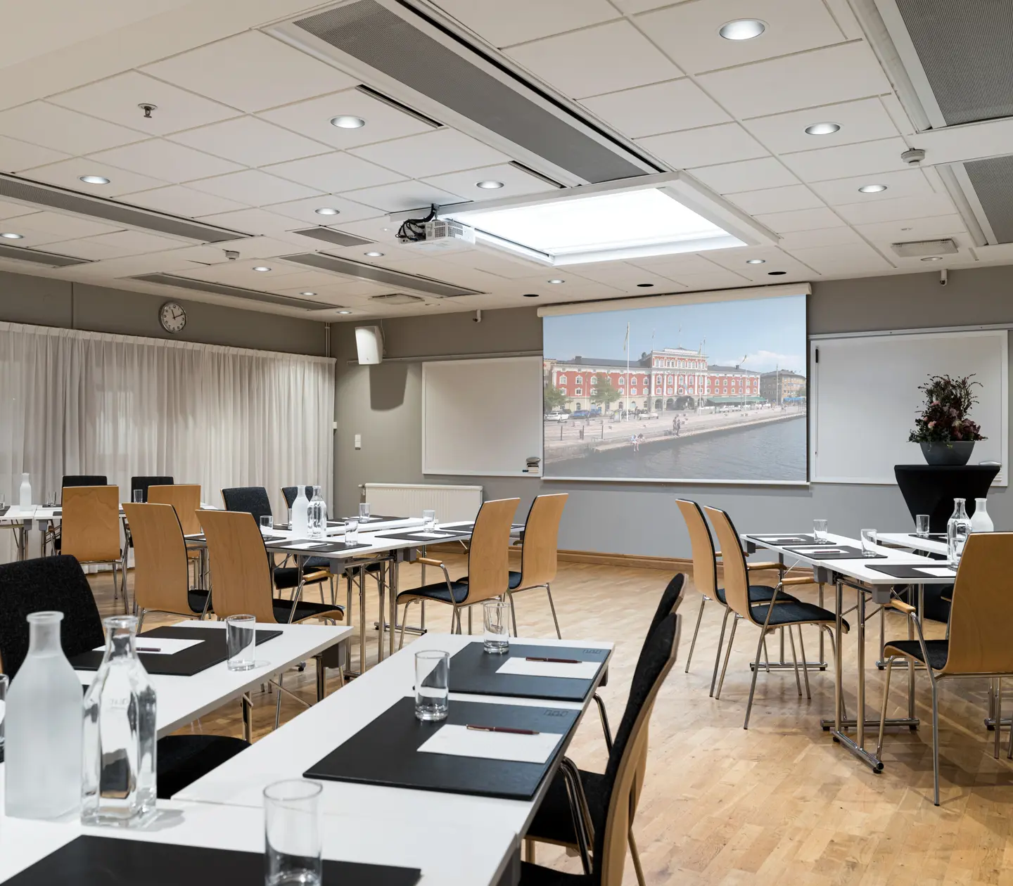 Spacious conference room setup with tables and chairs, featuring a large screen displaying a cityscape. The room has modern decor, a wooden floor, and is well-lit, ideal for meetings or presentations.