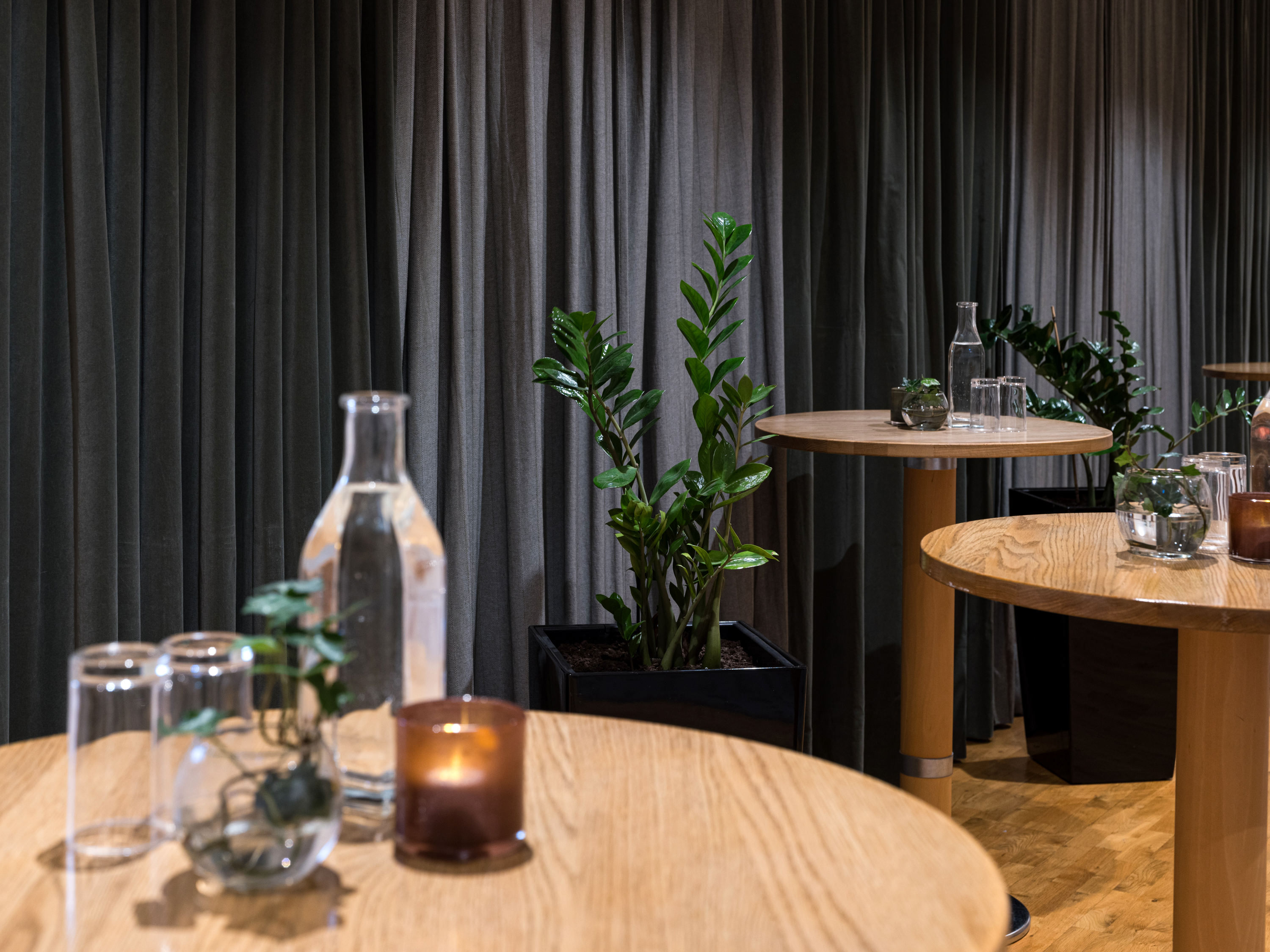 A modern interior space featuring wooden tables with glass bottles, candles, and small potted plants, surrounded by lush green plants and gray curtains in the background. Perfect for events or meetings with a natural decor theme.