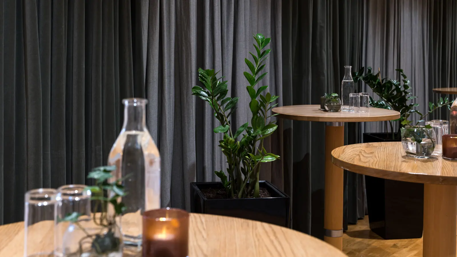A modern interior space featuring wooden tables with glass bottles, candles, and small potted plants, surrounded by lush green plants and gray curtains in the background. Perfect for events or meetings with a natural decor theme.