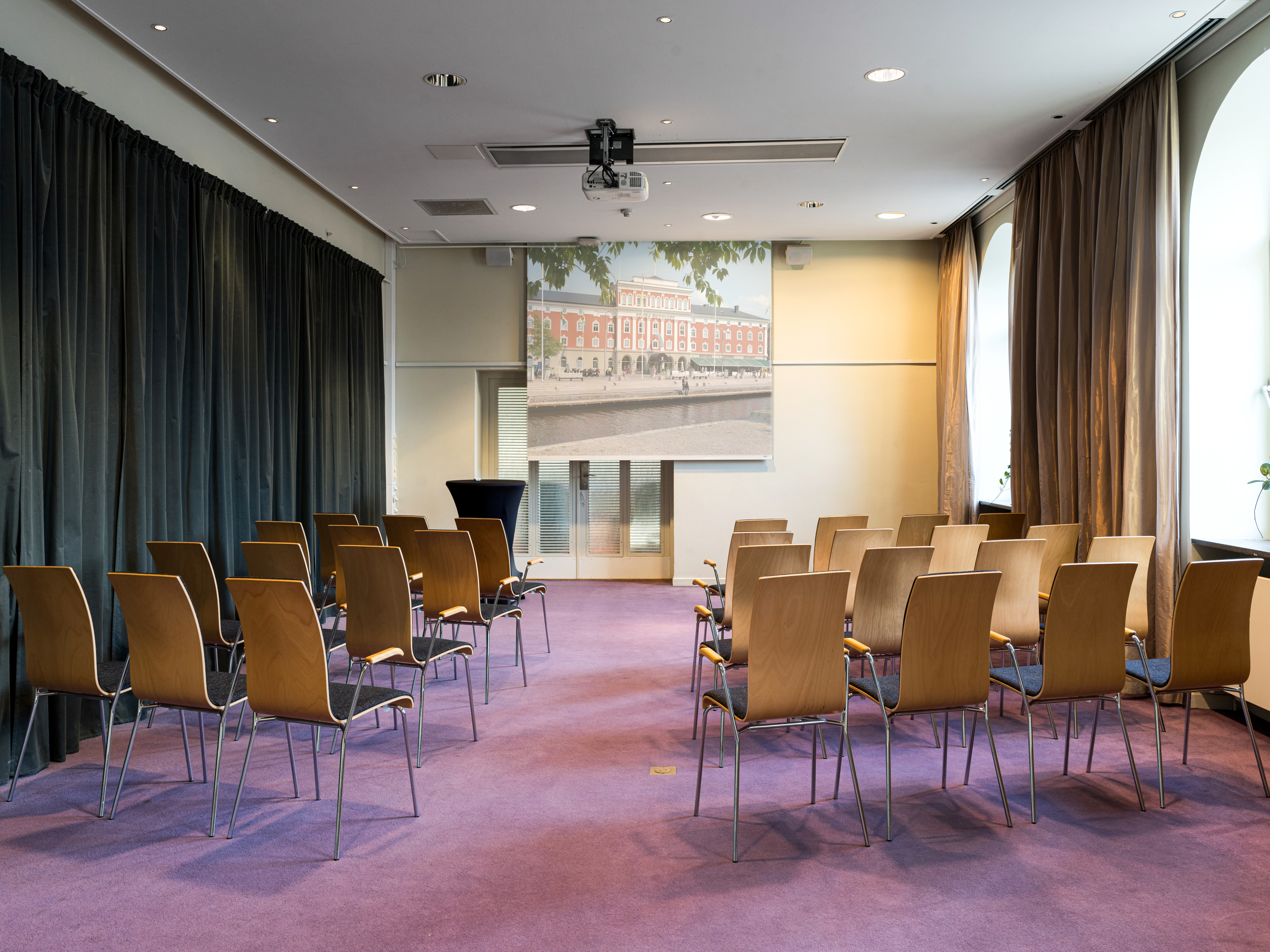 Conference room with arranged wooden chairs facing a large screen and projector, featuring natural light from tall windows and a soft purple carpet.