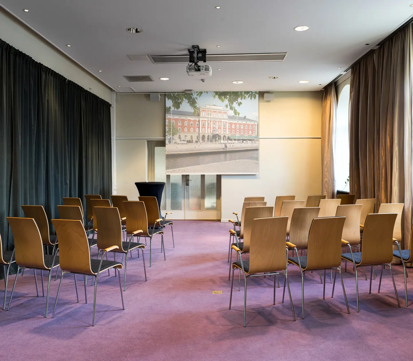 Conference room with arranged wooden chairs facing a large screen and projector, featuring natural light from tall windows and a soft purple carpet.