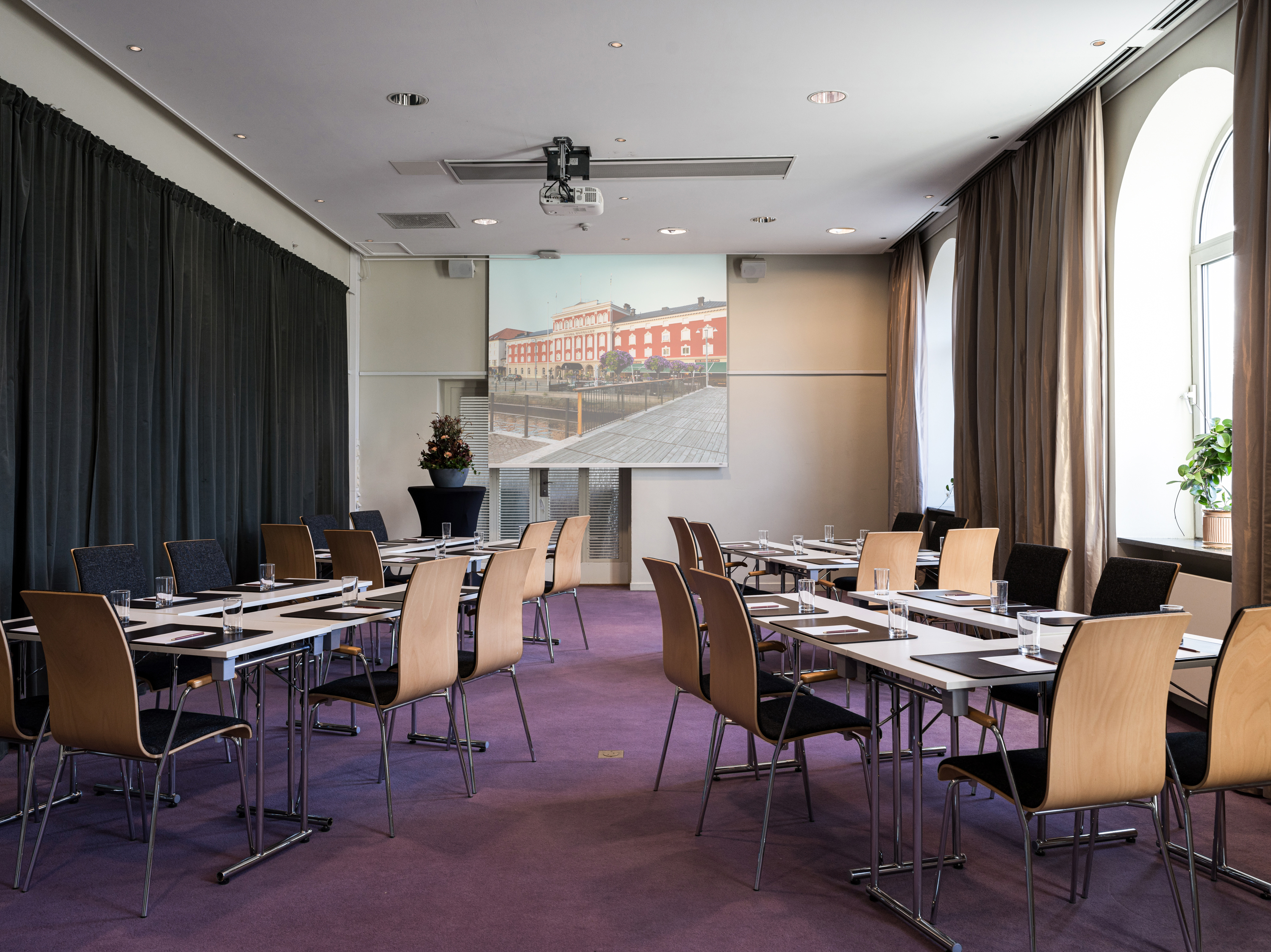 Modern conference room with rows of tables and chairs, featuring a large projector screen and natural light from arched windows.