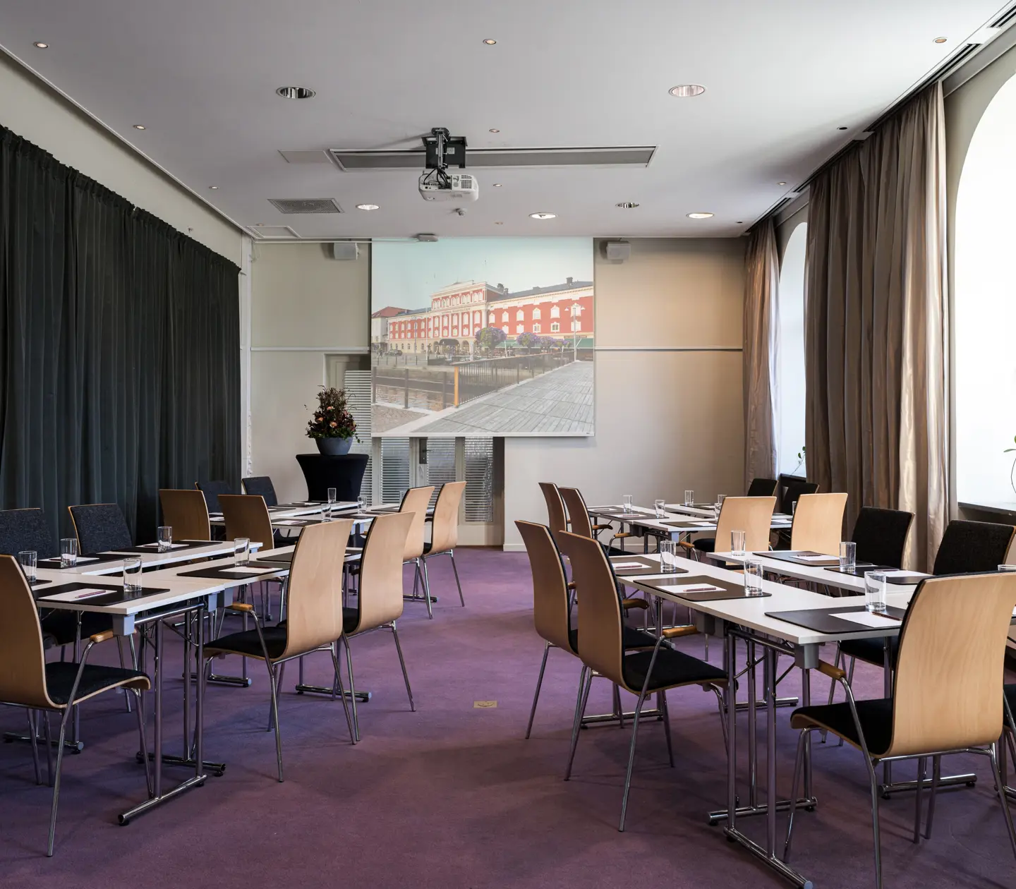 Modern conference room with rows of tables and chairs, featuring a large projector screen and natural light from arched windows.