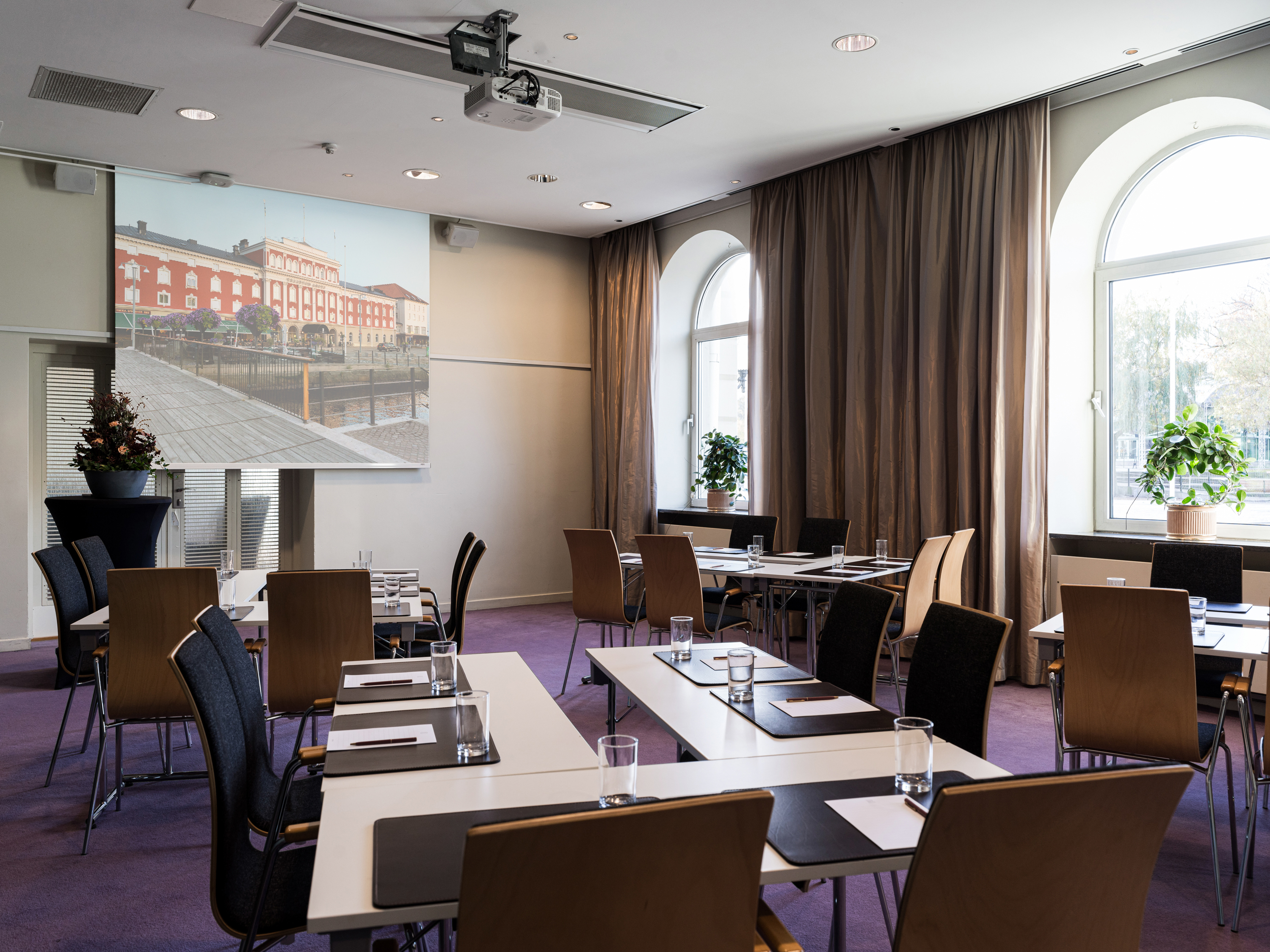 Modern conference room with arranged tables and chairs, featuring a large projection screen displaying a historic building. Bright natural light from arched windows and greenery create an inviting atmosphere. Ideal for meetings and presentations.