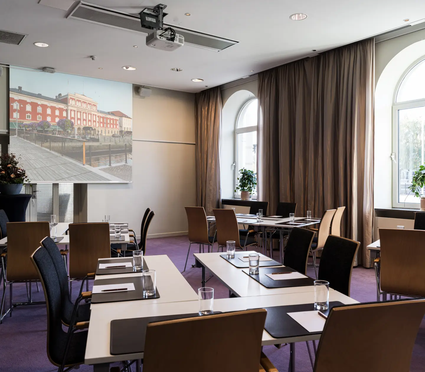 Modern conference room with arranged tables and chairs, featuring a large projection screen displaying a historic building. Bright natural light from arched windows and greenery create an inviting atmosphere. Ideal for meetings and presentations.