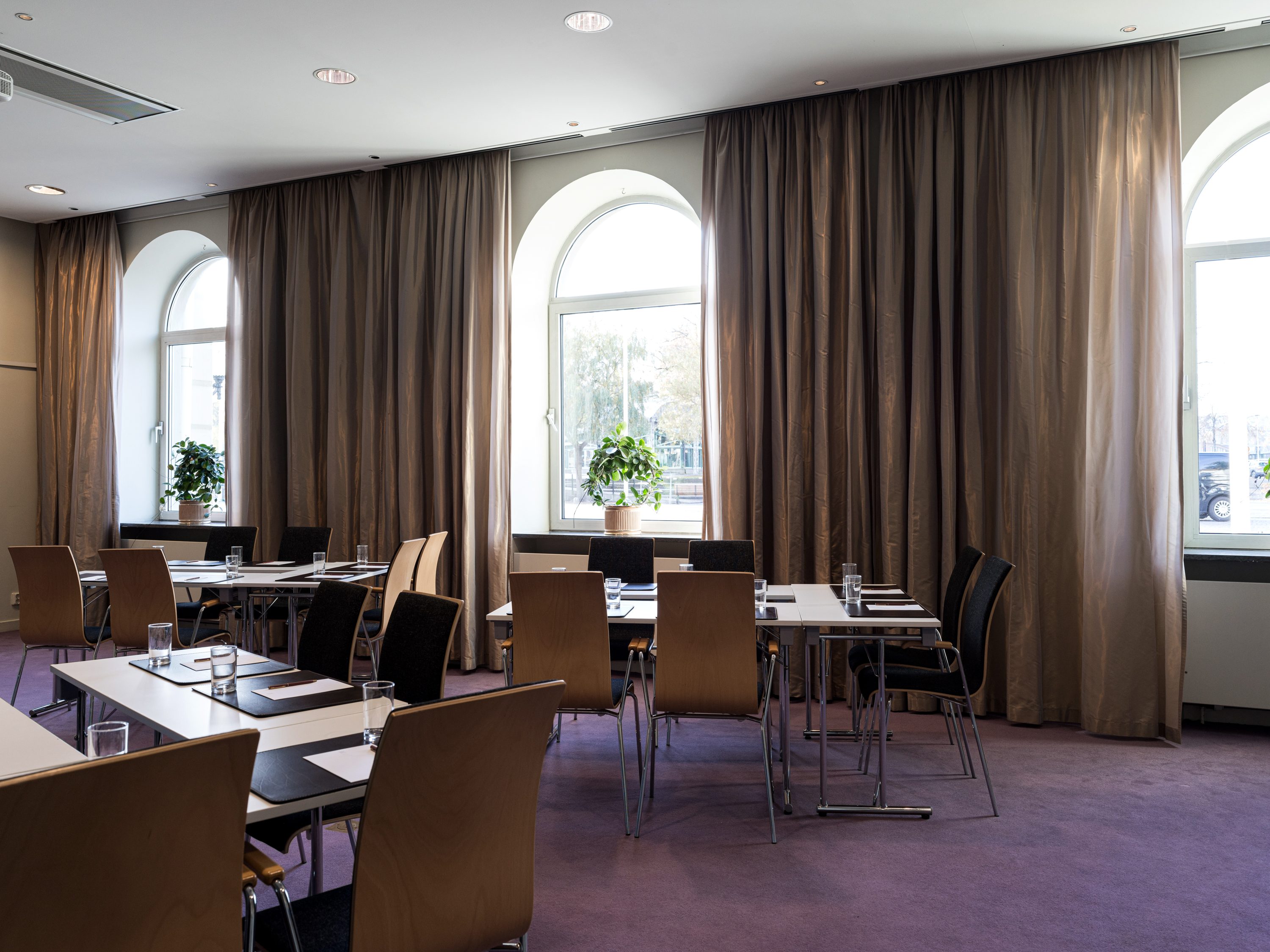 Modern conference room with wooden chairs, rectangular tables, and large windows with brown curtains, featuring indoor plants on window sills.