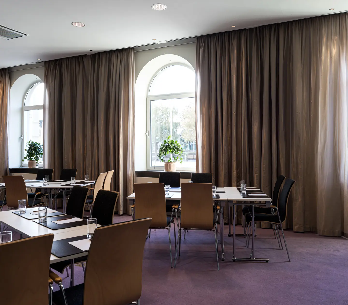 Modern conference room with wooden chairs, rectangular tables, and large windows with brown curtains, featuring indoor plants on window sills.