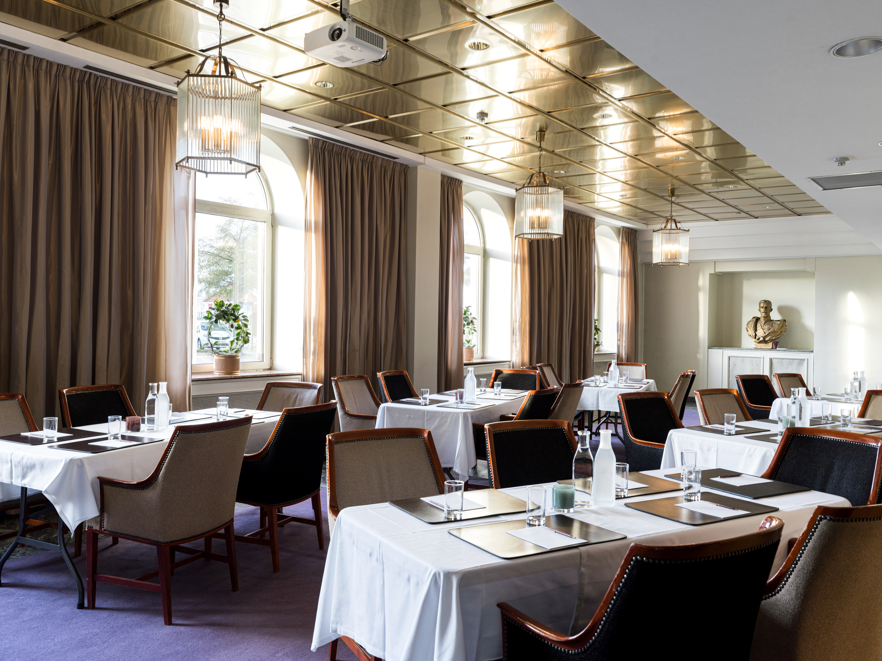 Elegant dining room with neatly arranged tables, white tablecloths, and a golden ceiling, featuring large windows with brown curtains and stylish pendant lights.