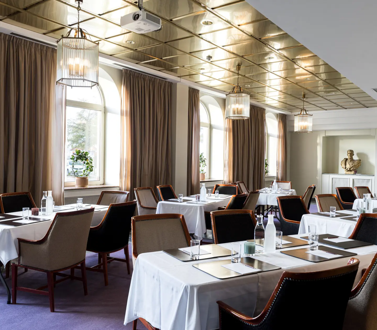 Elegant dining room with neatly arranged tables, white tablecloths, and a golden ceiling, featuring large windows with brown curtains and stylish pendant lights.