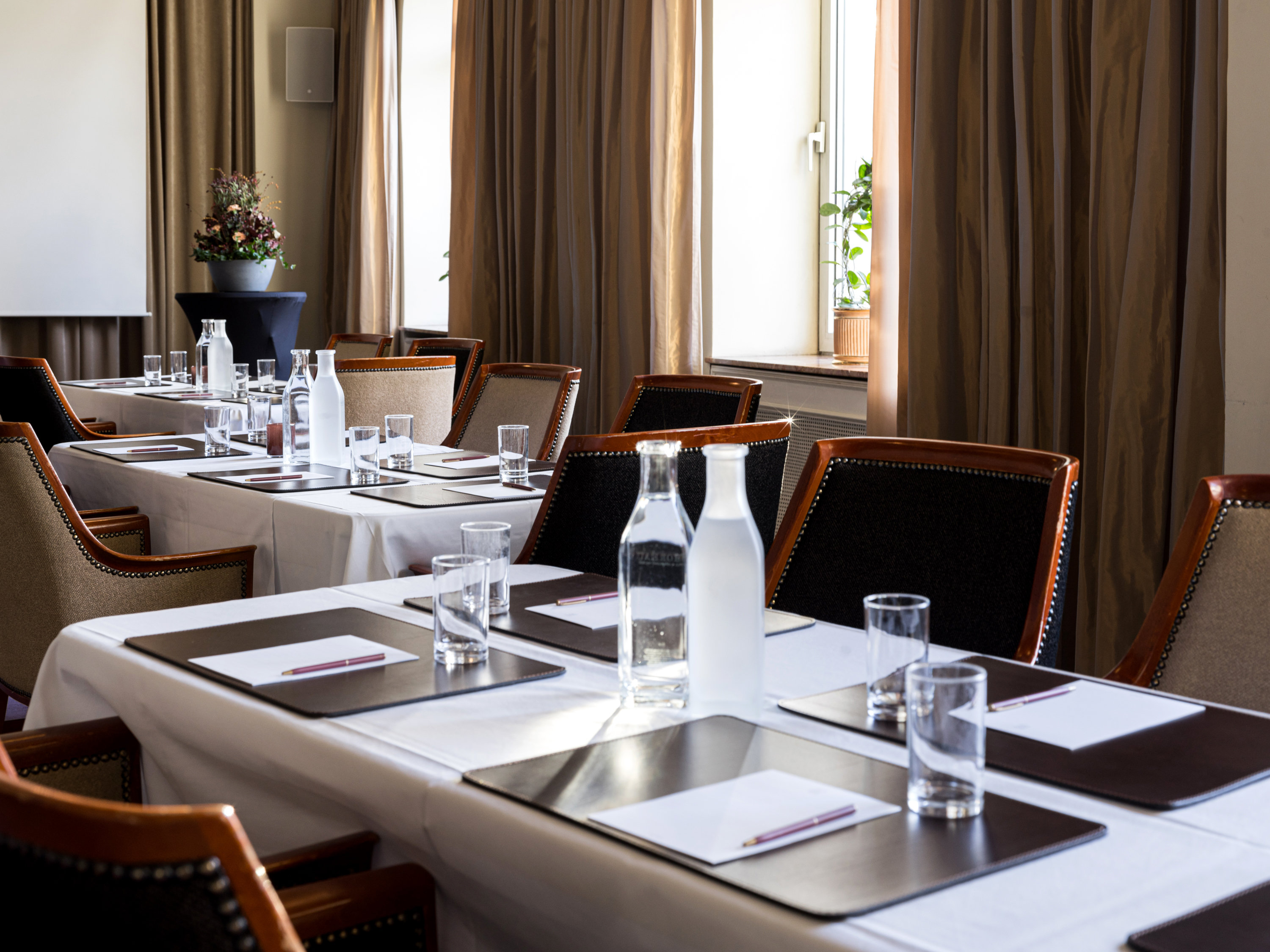 Elegant conference room setup with tables arranged for a meeting, featuring notebooks, pens, water bottles, and glasses, surrounded by comfortable cushioned chairs.