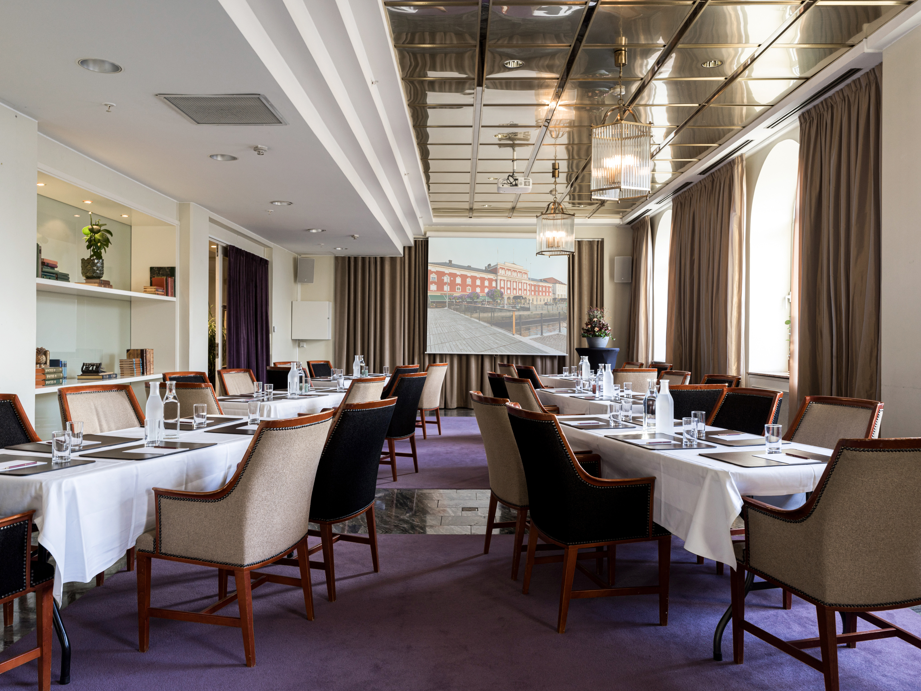 Elegant conference room with long tables, padded chairs, and a projector screen displaying a cityscape. The room features modern decor with large windows and purple carpet, ideal for meetings and presentations.