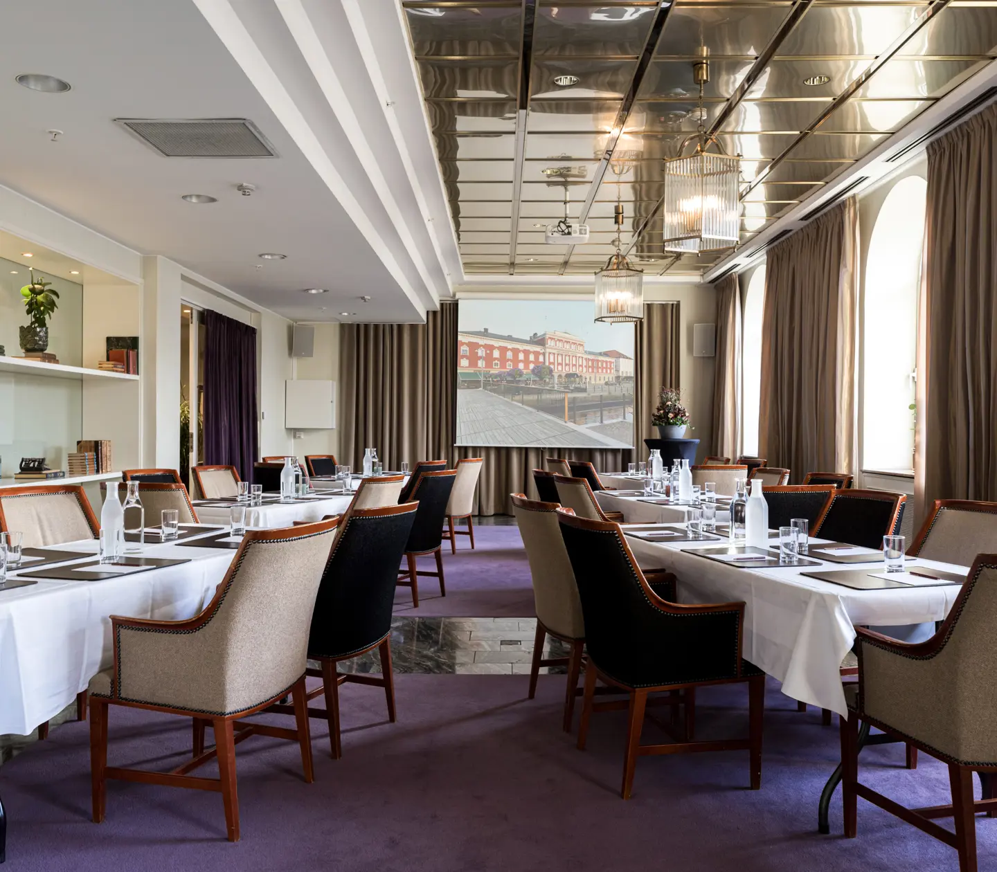 Elegant conference room with long tables, padded chairs, and a projector screen displaying a cityscape. The room features modern decor with large windows and purple carpet, ideal for meetings and presentations.