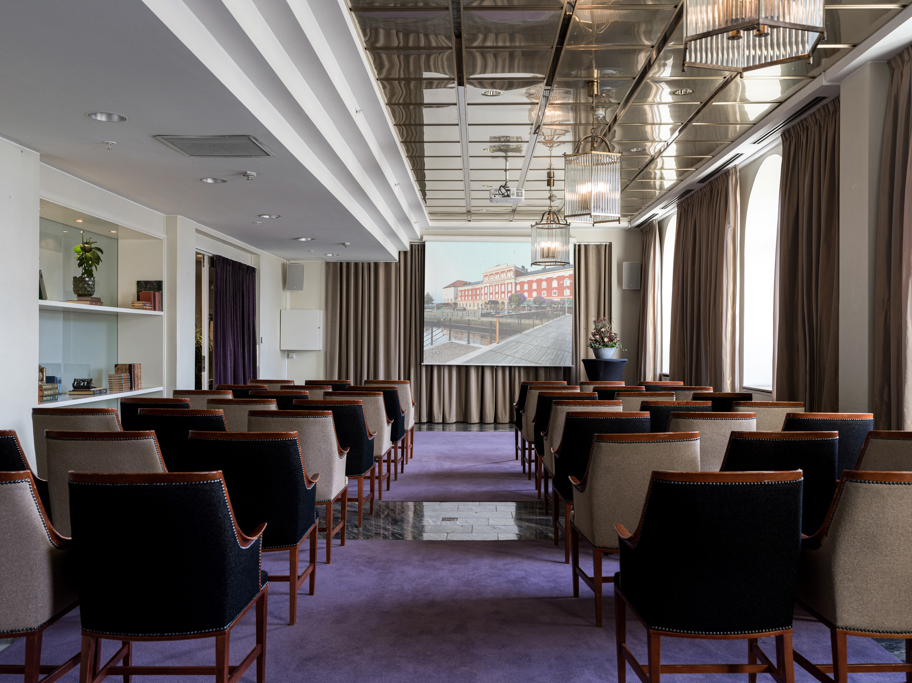 Conference room with rows of upholstered chairs facing a large screen, featuring modern lighting and large windows with curtains, creating a professional setting.