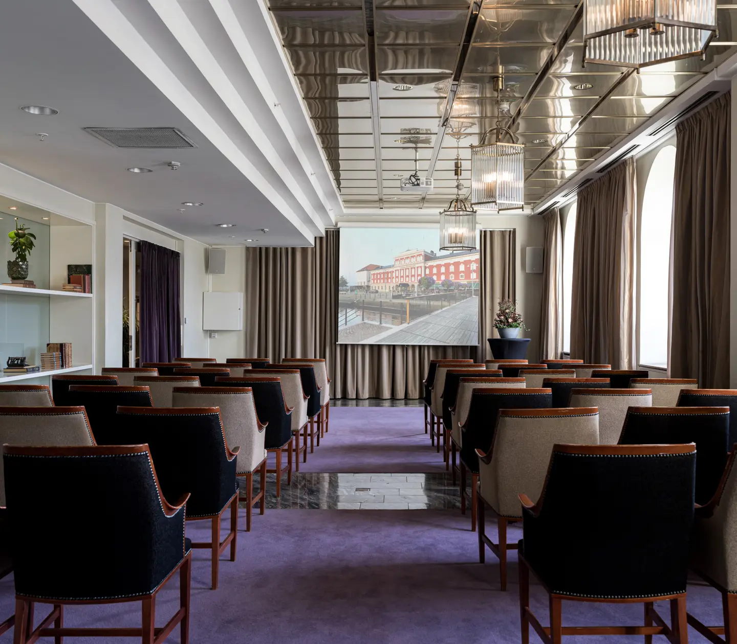 Conference room with rows of upholstered chairs facing a large screen, featuring modern lighting and large windows with curtains, creating a professional setting.