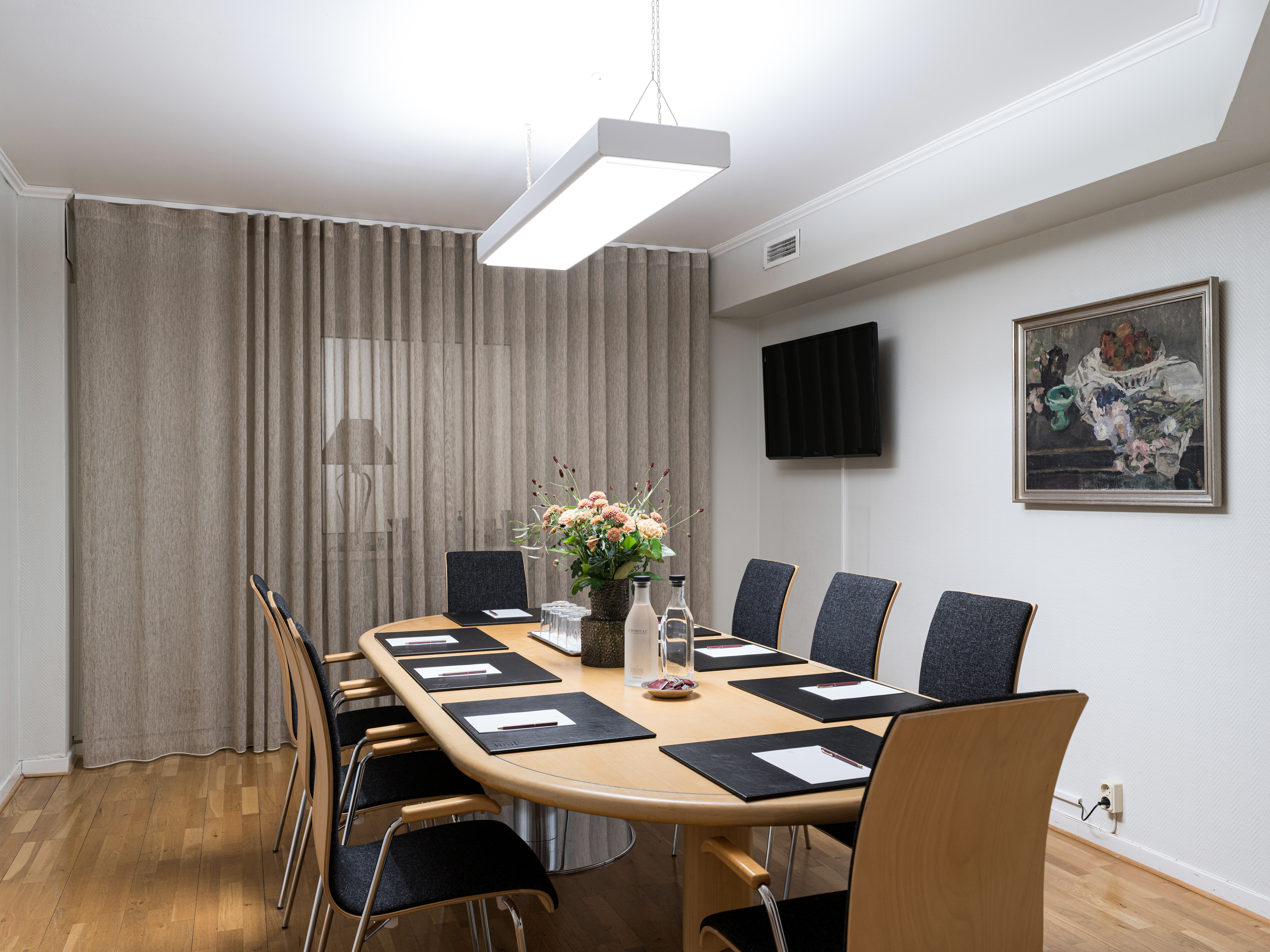 Modern conference room with a wooden table, black chairs, notepads, and a vase of flowers. The room is well-lit with a ceiling light, decorative painting, and wall-mounted TV. Neutral curtains cover one wall.