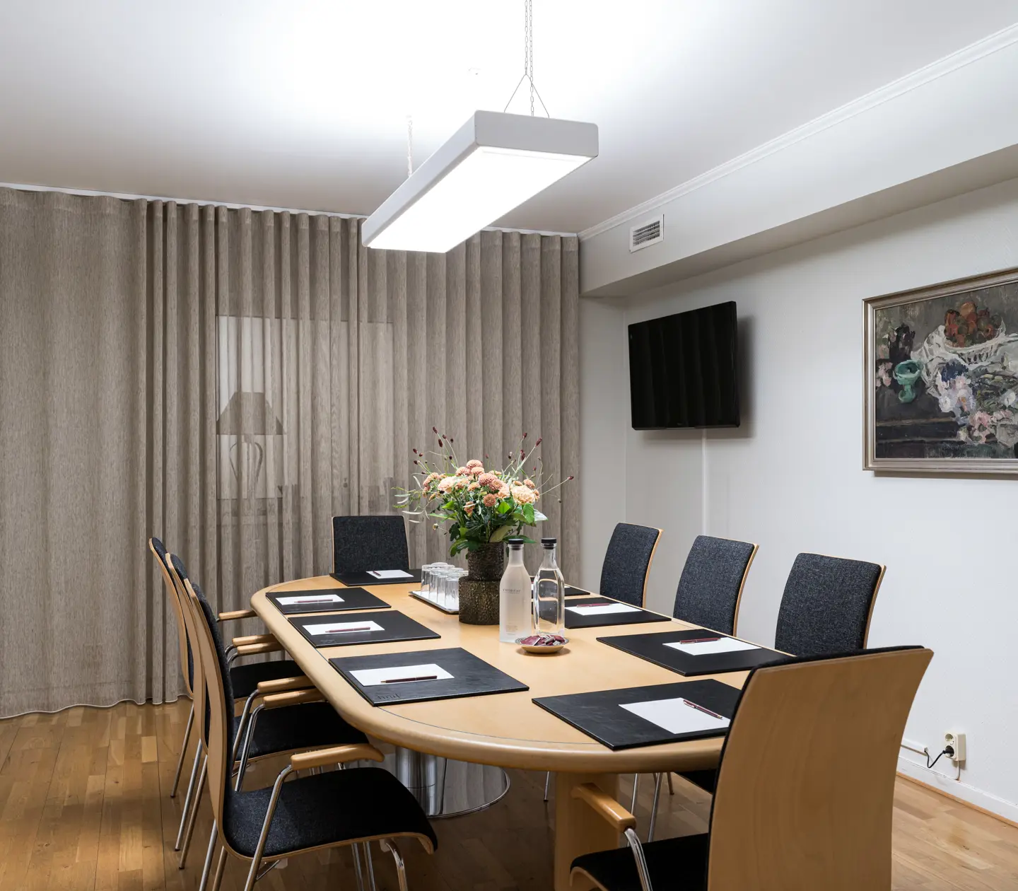 Modern conference room with a wooden table, black chairs, notepads, and a vase of flowers. The room is well-lit with a ceiling light, decorative painting, and wall-mounted TV. Neutral curtains cover one wall.