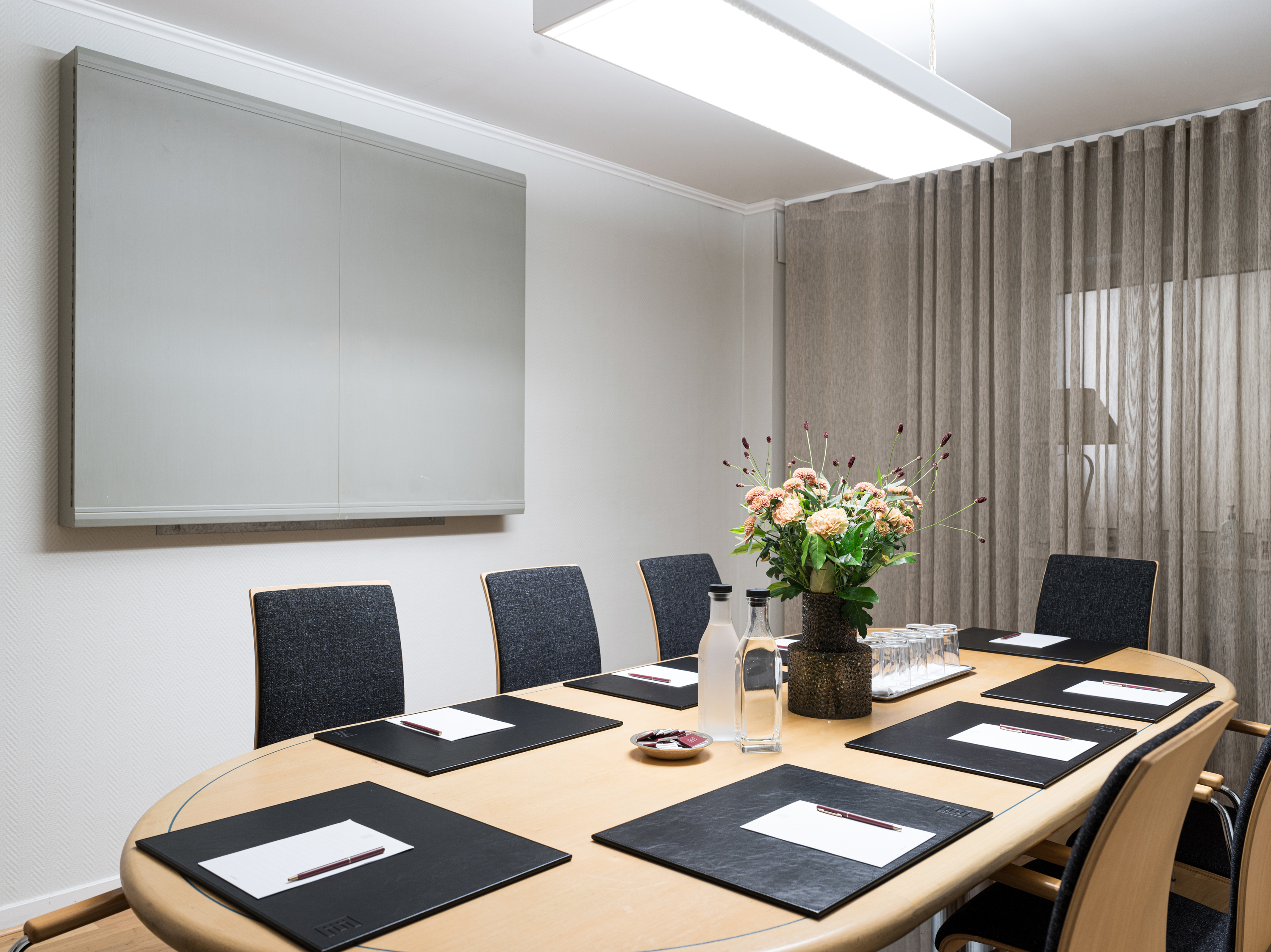 Modern conference room with a wooden oval table, surrounded by black chairs, featuring notepads, pens, water bottles, and a floral centerpiece, set against a backdrop of white walls and sheer curtains.
