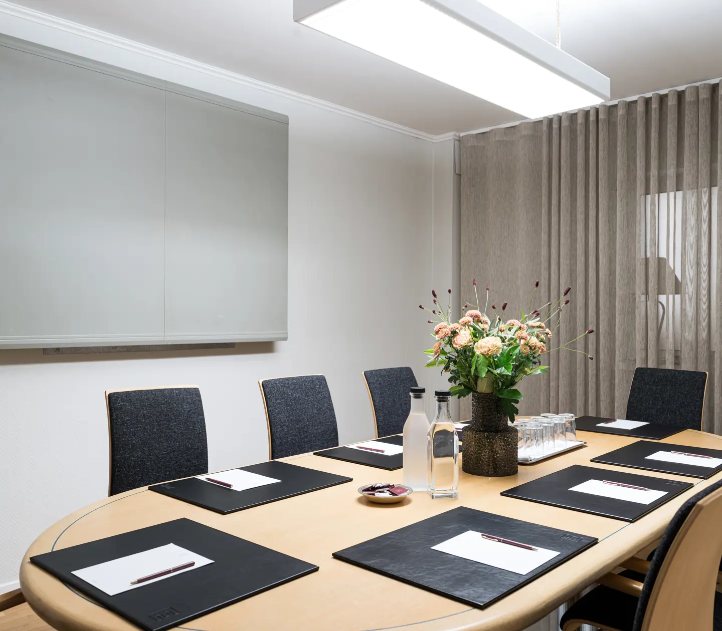 Modern conference room with a wooden oval table, surrounded by black chairs, featuring notepads, pens, water bottles, and a floral centerpiece, set against a backdrop of white walls and sheer curtains.