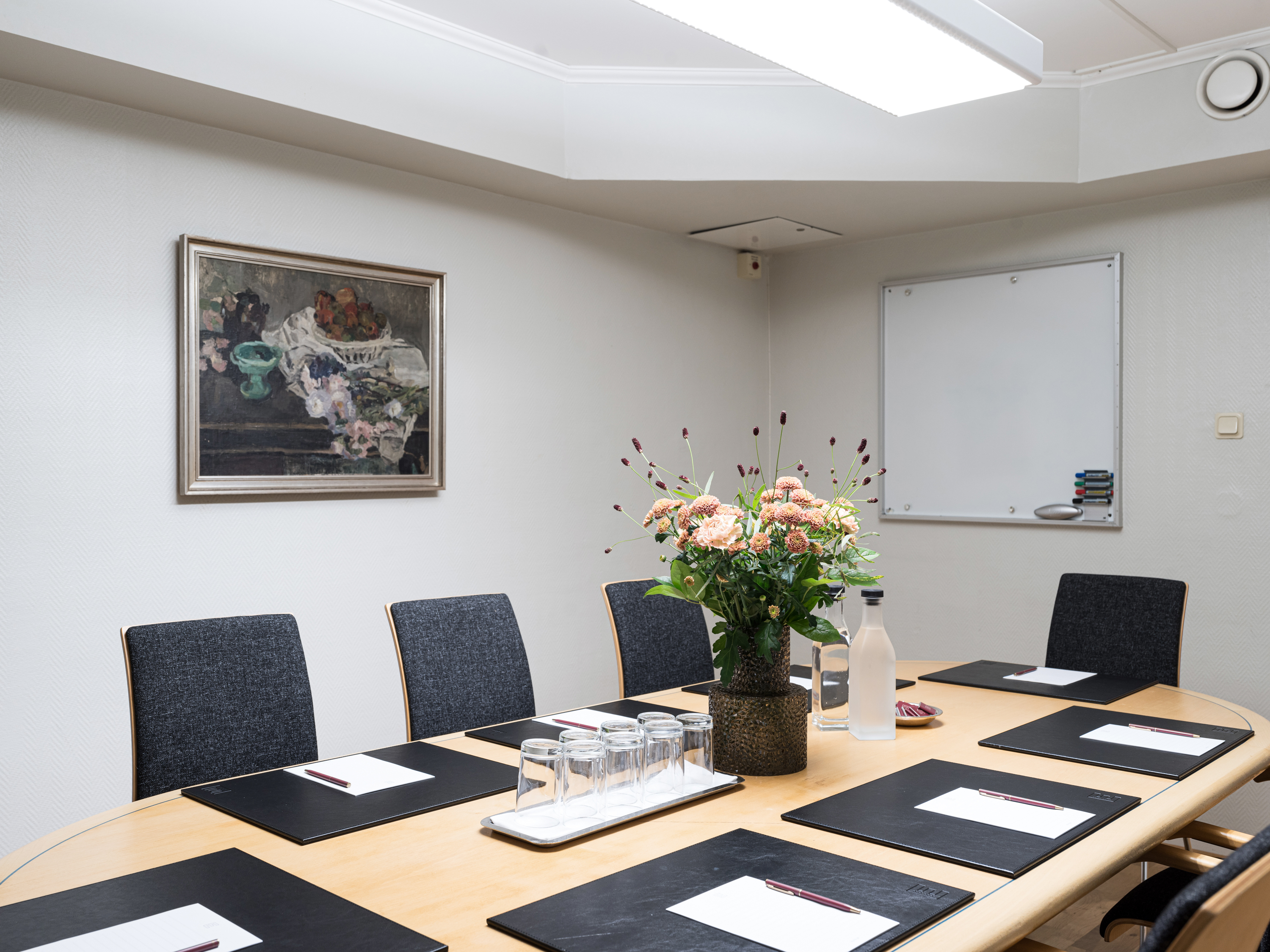 Modern conference room with oval table, six chairs, notepads, pens, a bouquet of flowers centerpiece, and a whiteboard.