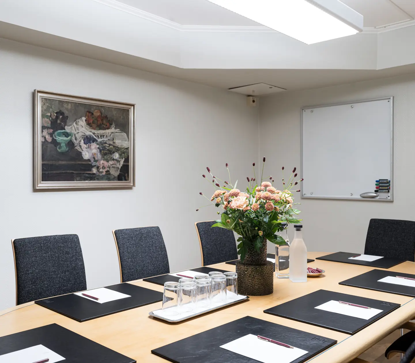 Modern conference room with oval table, six chairs, notepads, pens, a bouquet of flowers centerpiece, and a whiteboard.