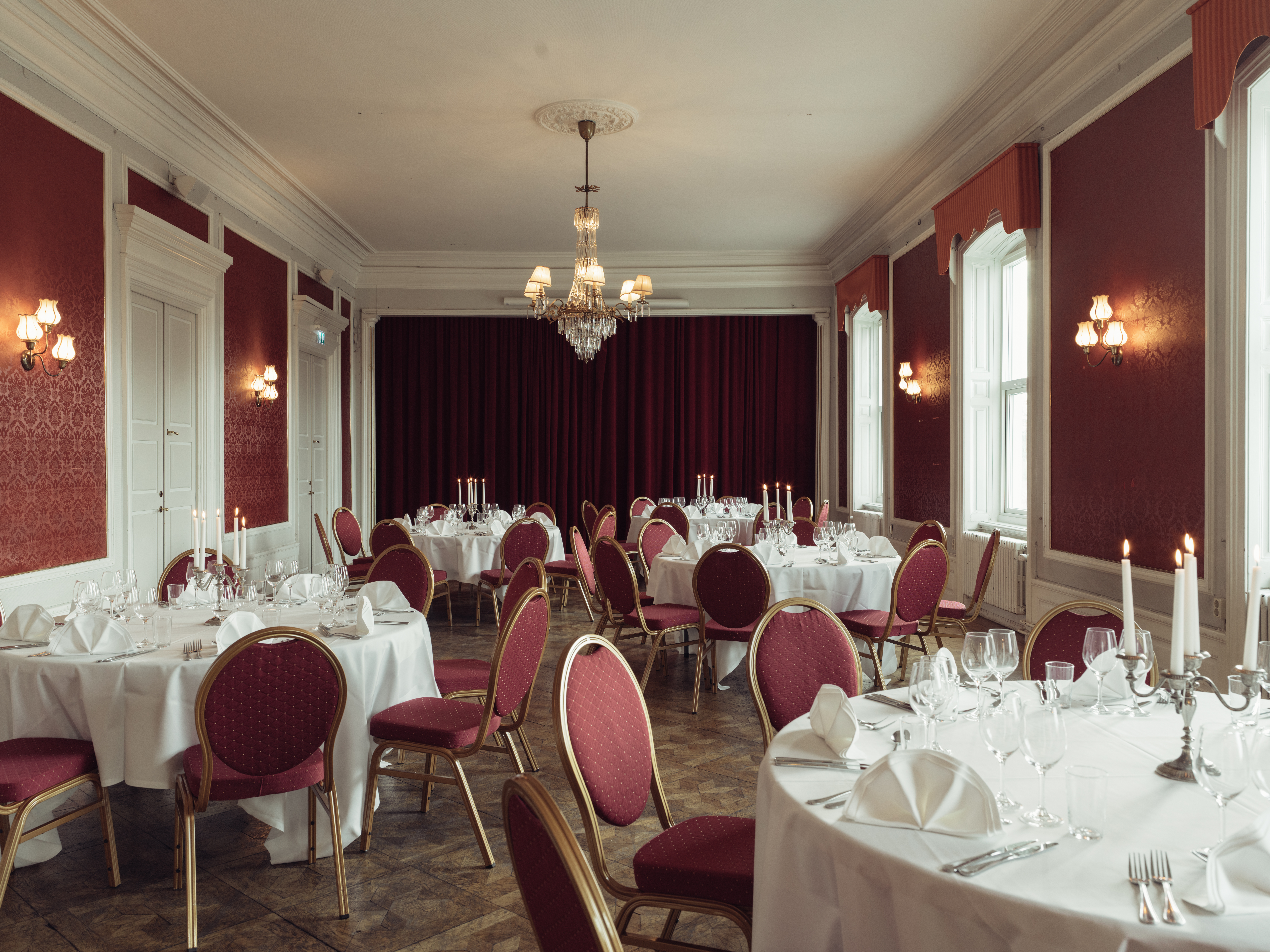 Elegant banquet hall with round tables draped in white tablecloths, red chairs, and a chandelier, set up for a formal event.