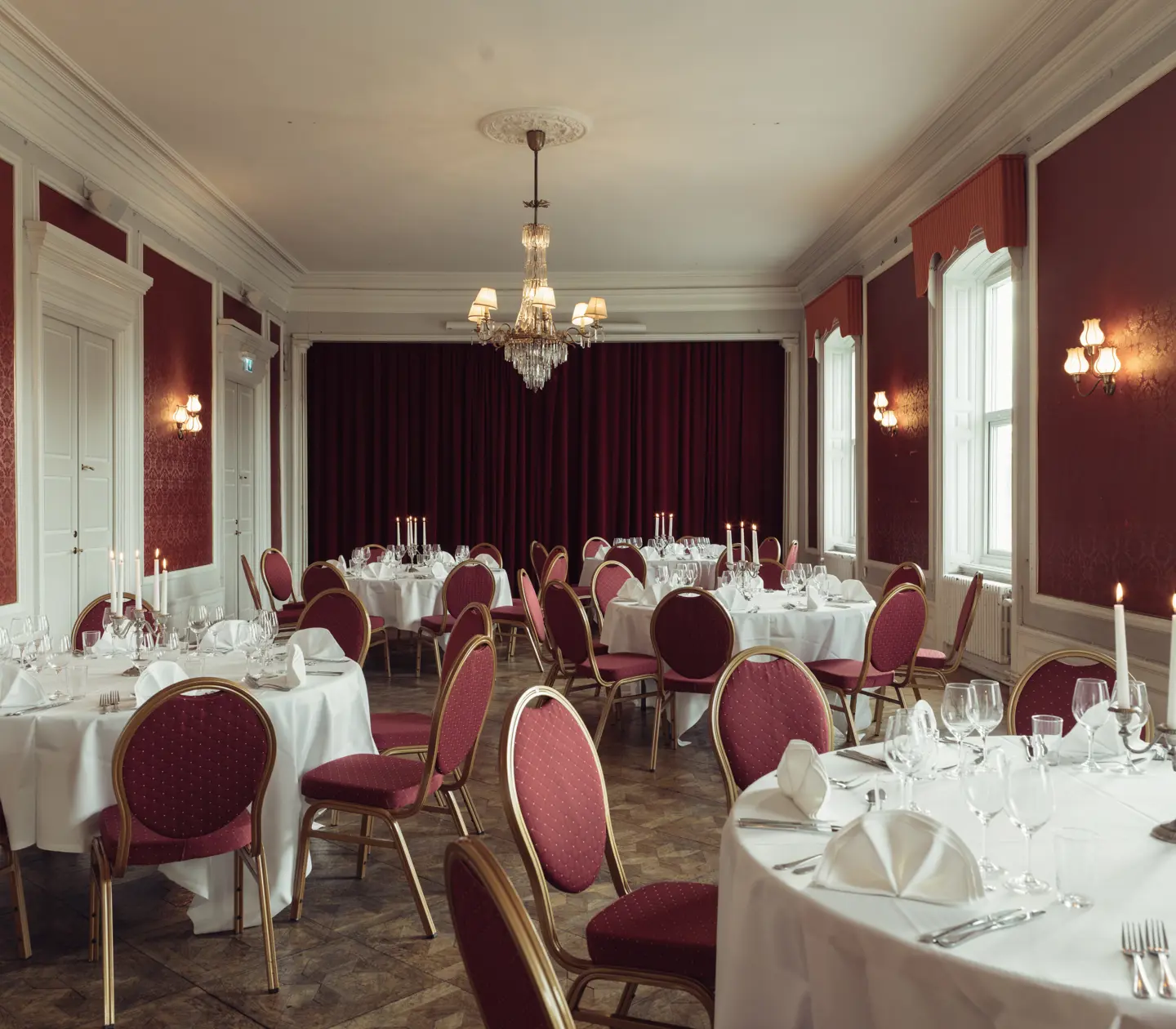 Elegant banquet hall with round tables draped in white tablecloths, red chairs, and a chandelier, set up for a formal event.