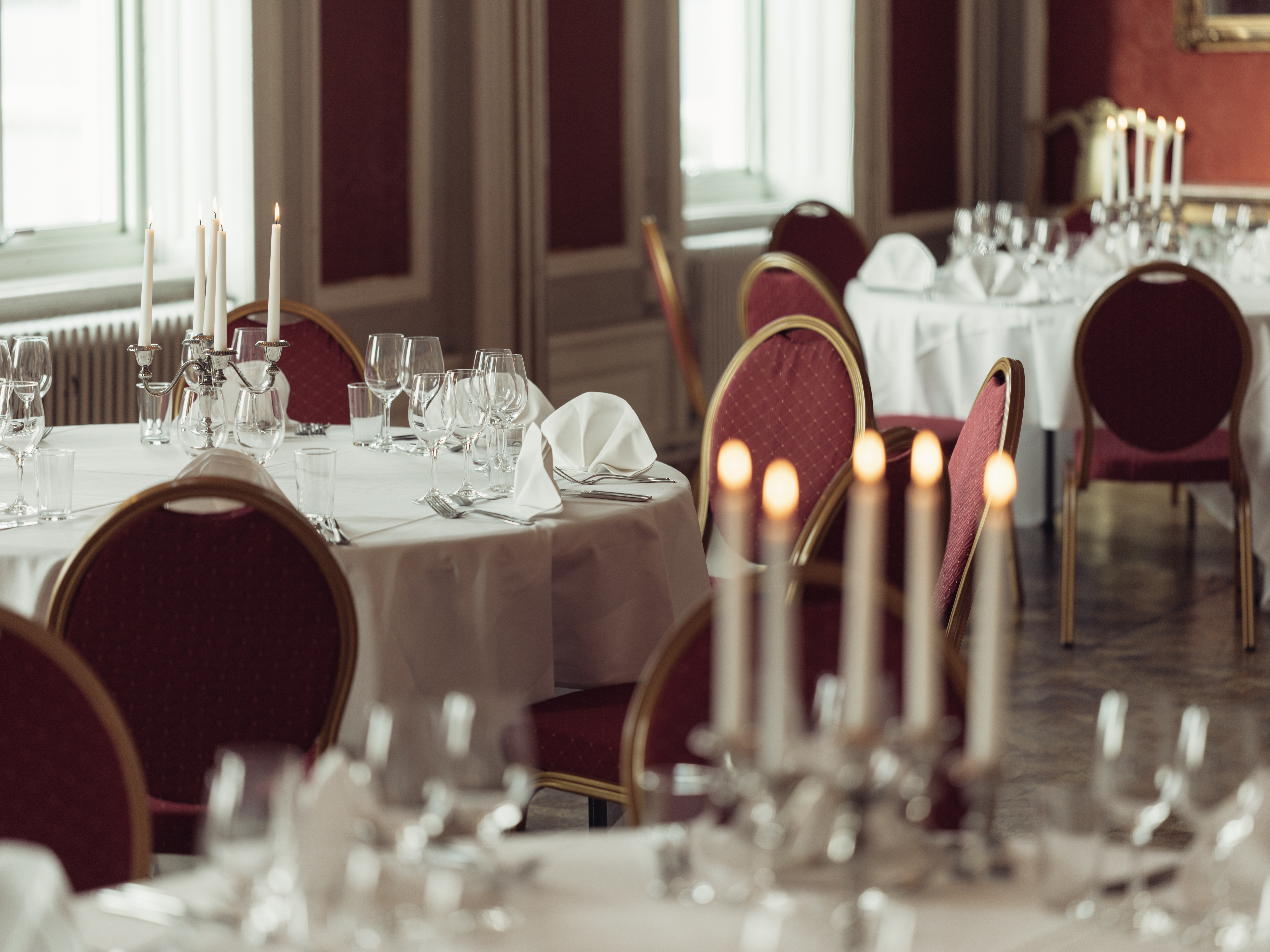 Elegant restaurant dining room with round tables set for an event, featuring white tablecloths, red chairs, folded napkins, candles, and neatly arranged glassware.