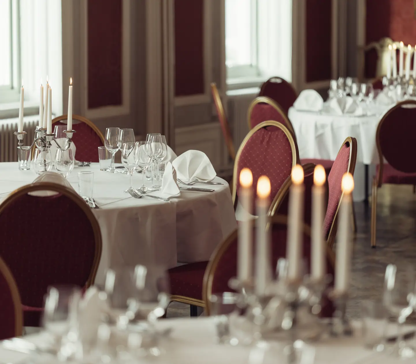 Elegant restaurant dining room with round tables set for an event, featuring white tablecloths, red chairs, folded napkins, candles, and neatly arranged glassware.