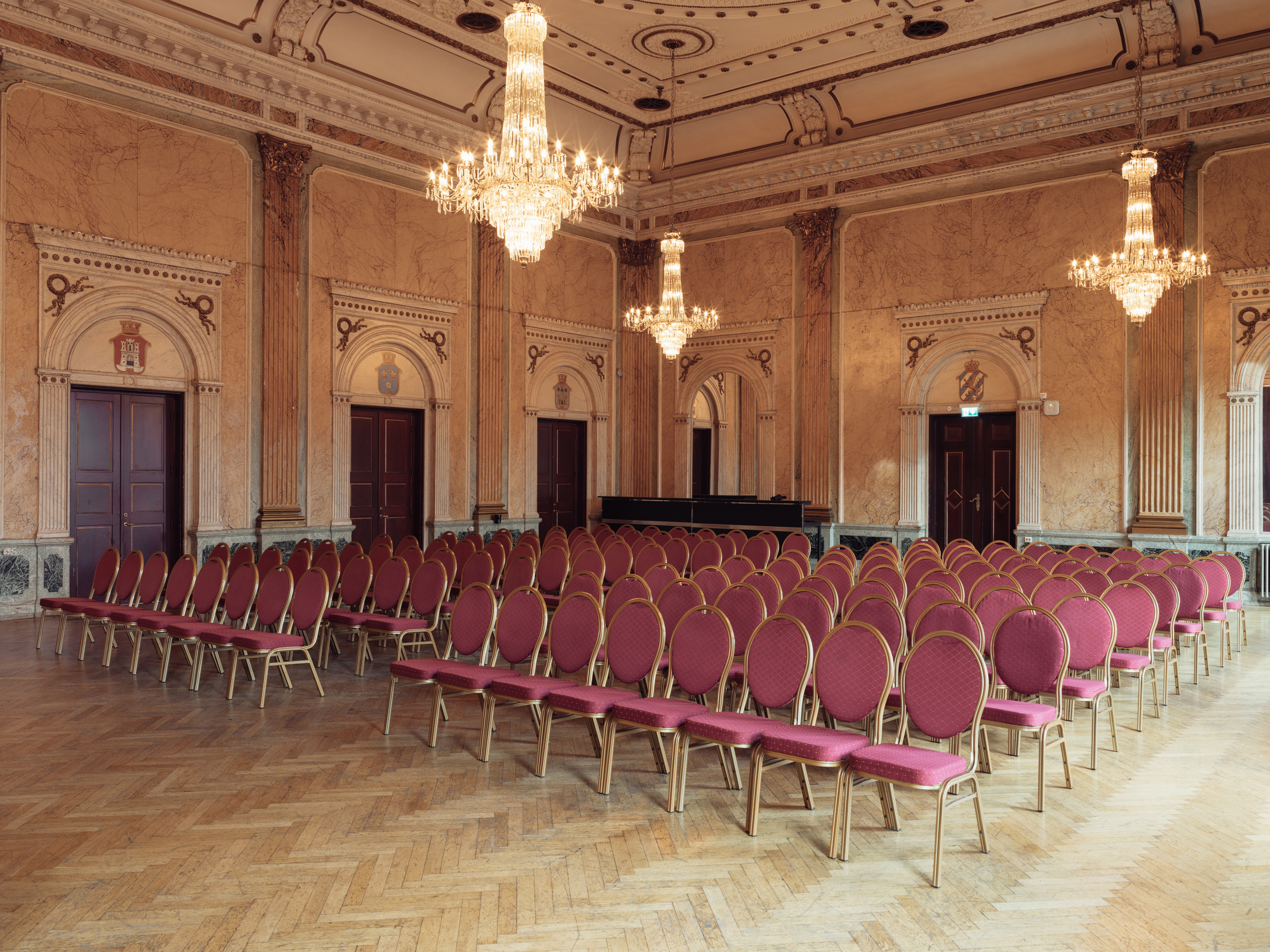 Elegant banquet hall with historic architecture. The room features beautiful wall panels and decorated doors, with large crystal chandeliers hanging from the ceiling. Rows of chairs with red upholstery and golden frames are arranged on a herringbone-patterned wooden