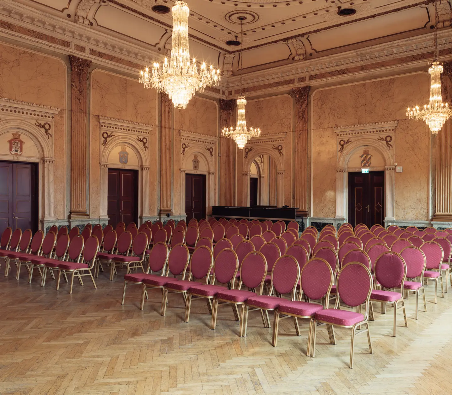 Elegant banquet hall with historic architecture. The room features beautiful wall panels and decorated doors, with large crystal chandeliers hanging from the ceiling. Rows of chairs with red upholstery and golden frames are arranged on a herringbone-patterned wooden