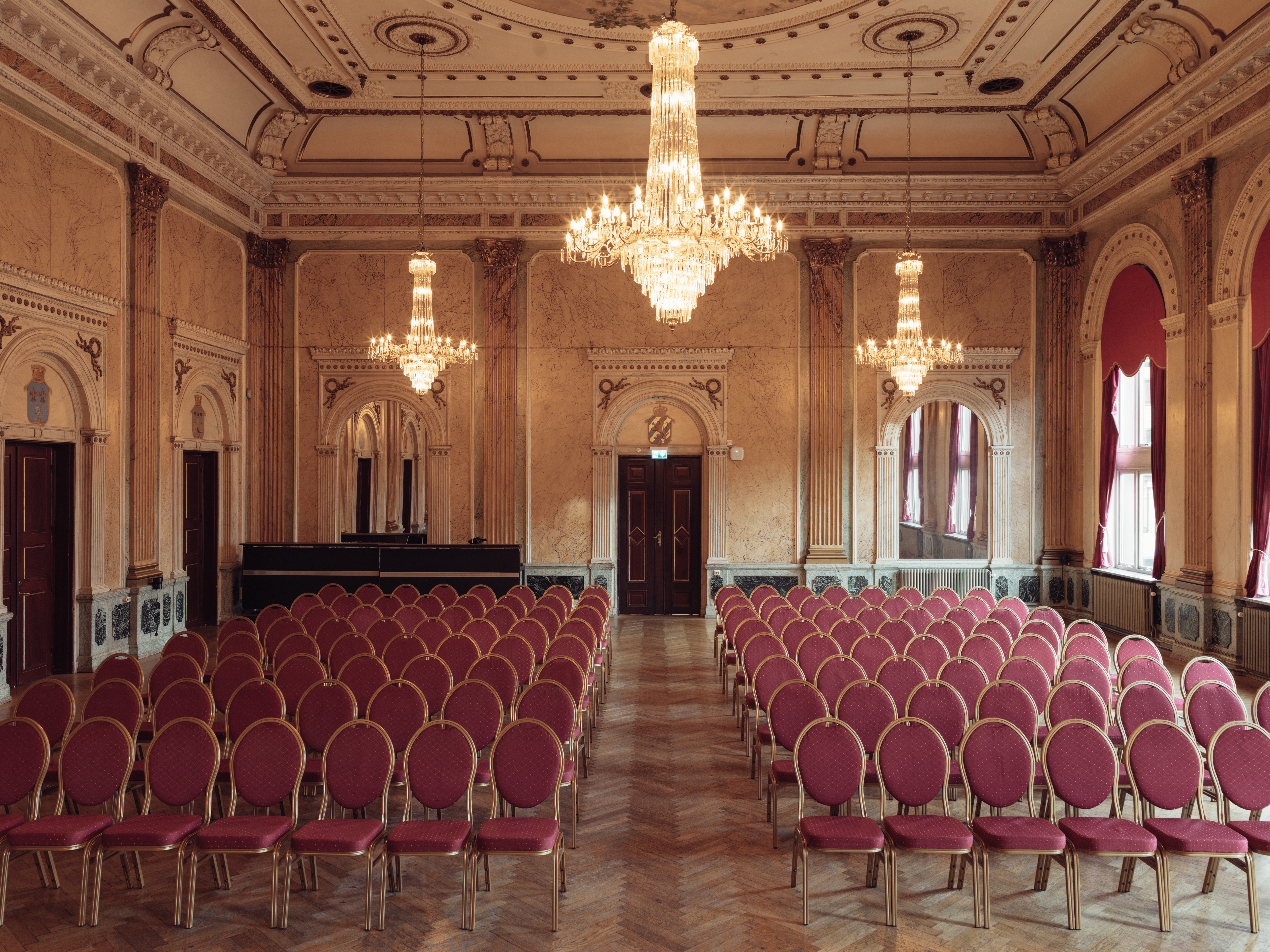 Elegant ballroom with vintage chandeliers, red velvet chairs, and ornate walls, set for an upscale event.
