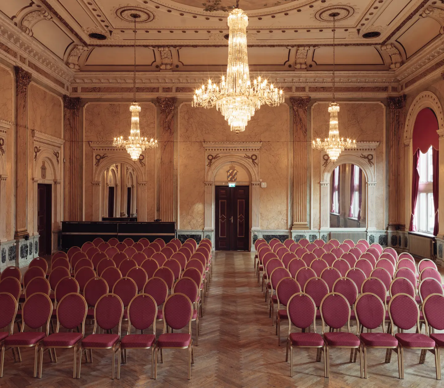 Elegant ballroom with vintage chandeliers, red velvet chairs, and ornate walls, set for an upscale event.