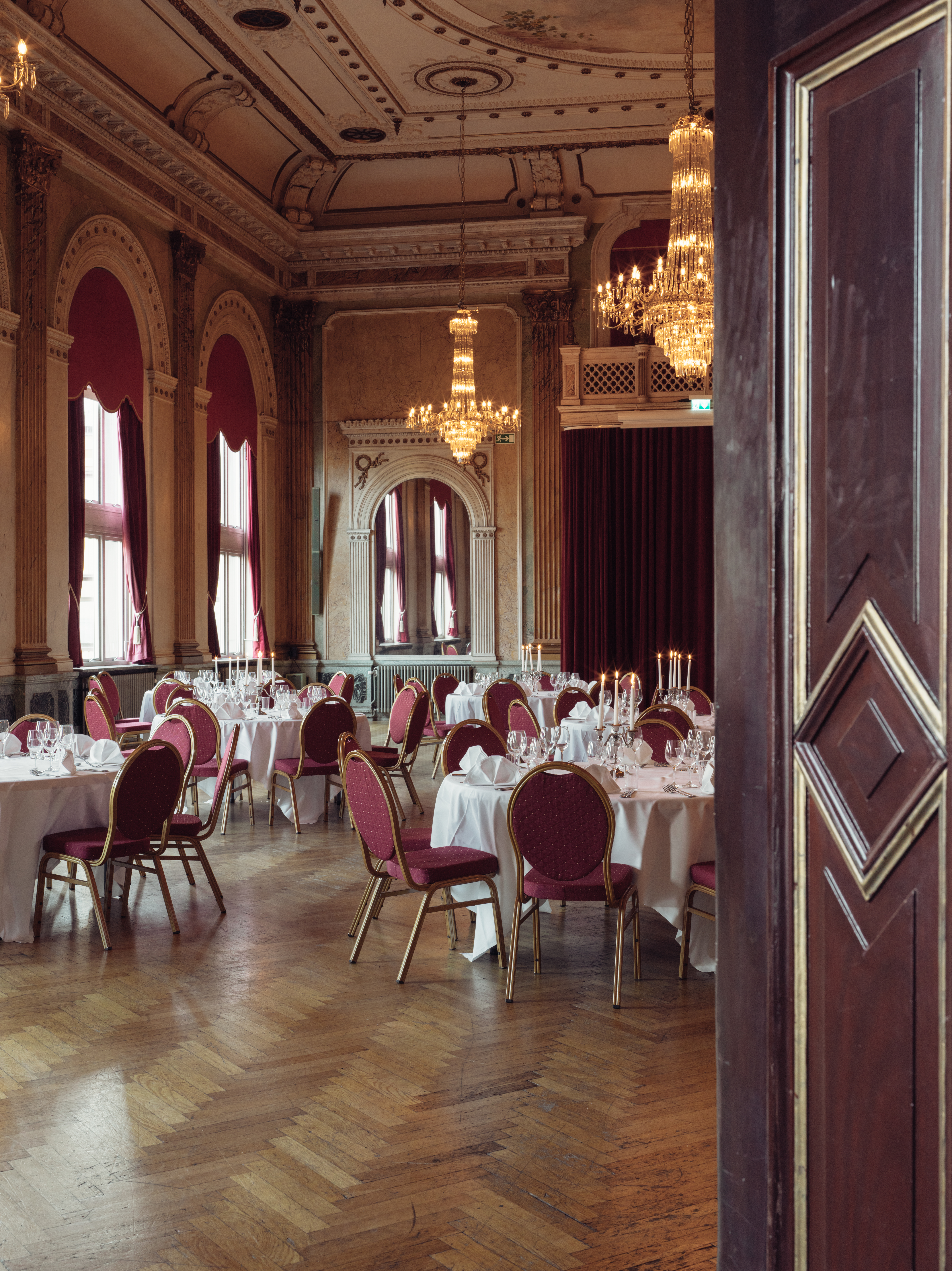 Elegant banquet hall with round tables, draped in white tablecloths, set for an event. The classic decor features arched windows with red curtains, ornate chandeliers, and wooden floors, creating a luxurious and timeless atmosphere.