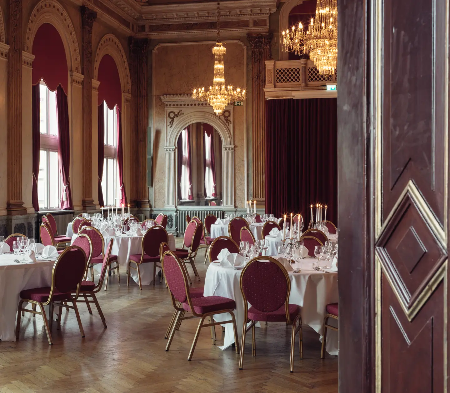 Elegant banquet hall with round tables, draped in white tablecloths, set for an event. The classic decor features arched windows with red curtains, ornate chandeliers, and wooden floors, creating a luxurious and timeless atmosphere.