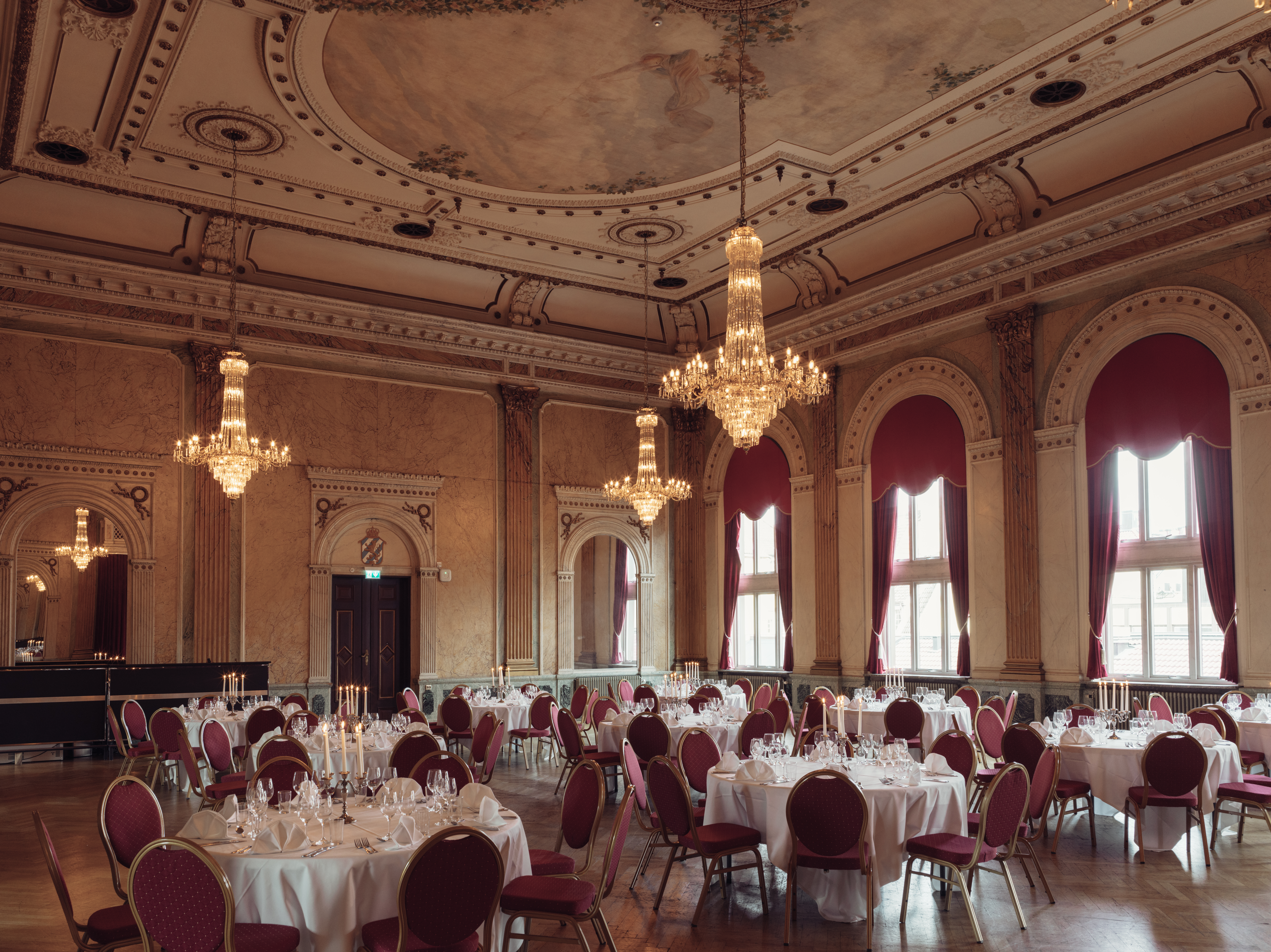 Elegant ballroom with ornate chandeliers, red draped windows, and round tables set for a formal event.