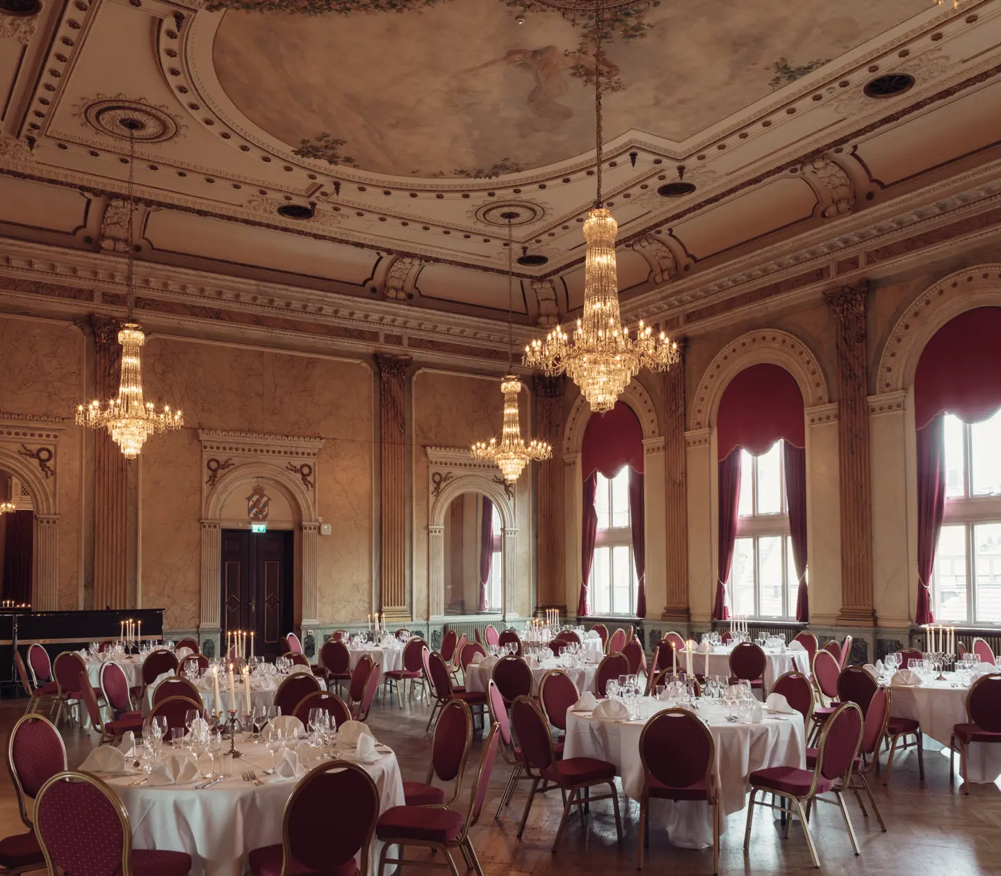 Elegant ballroom with ornate chandeliers, red draped windows, and round tables set for a formal event.